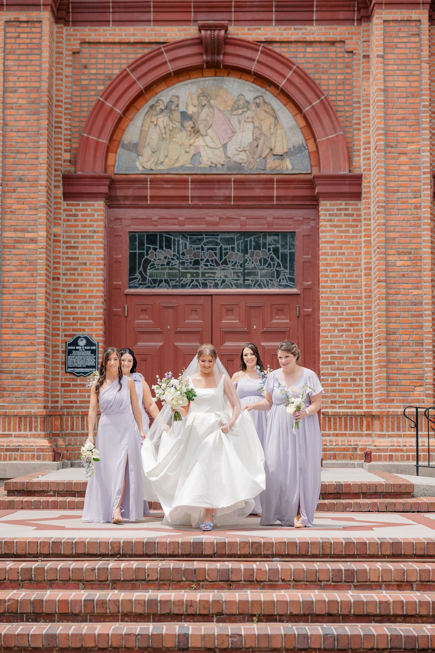 Bride in white gown with four bridesmaids in lavender dresses on brick church steps.