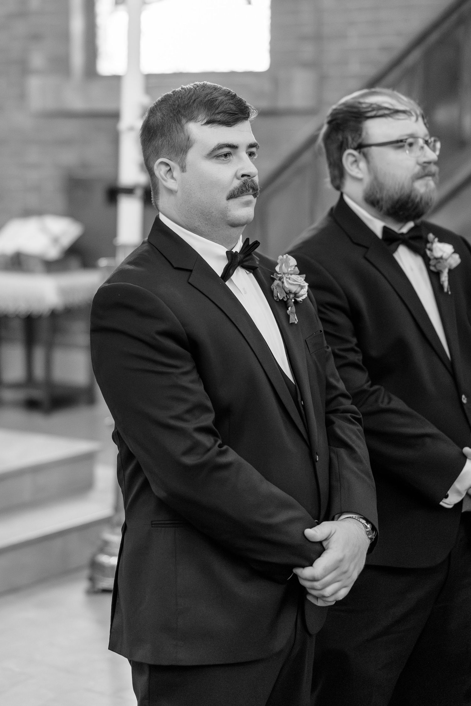 Groom in black tuxedo with boutonniere and bow tie stands at altar awaiting bride during wedding ceremony.