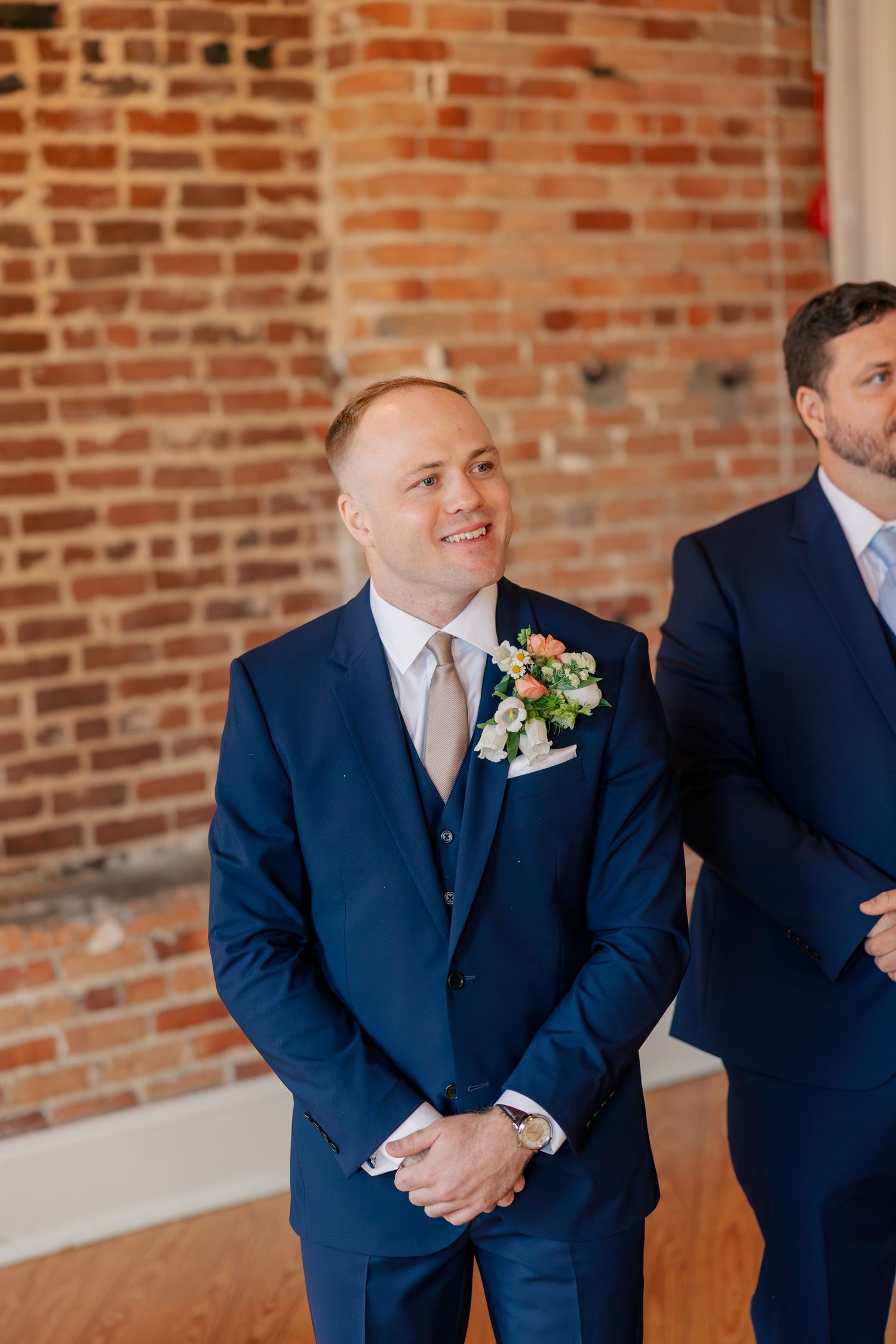Handsome groom in navy three-piece suit with floral boutonniere smiling during ceremony.