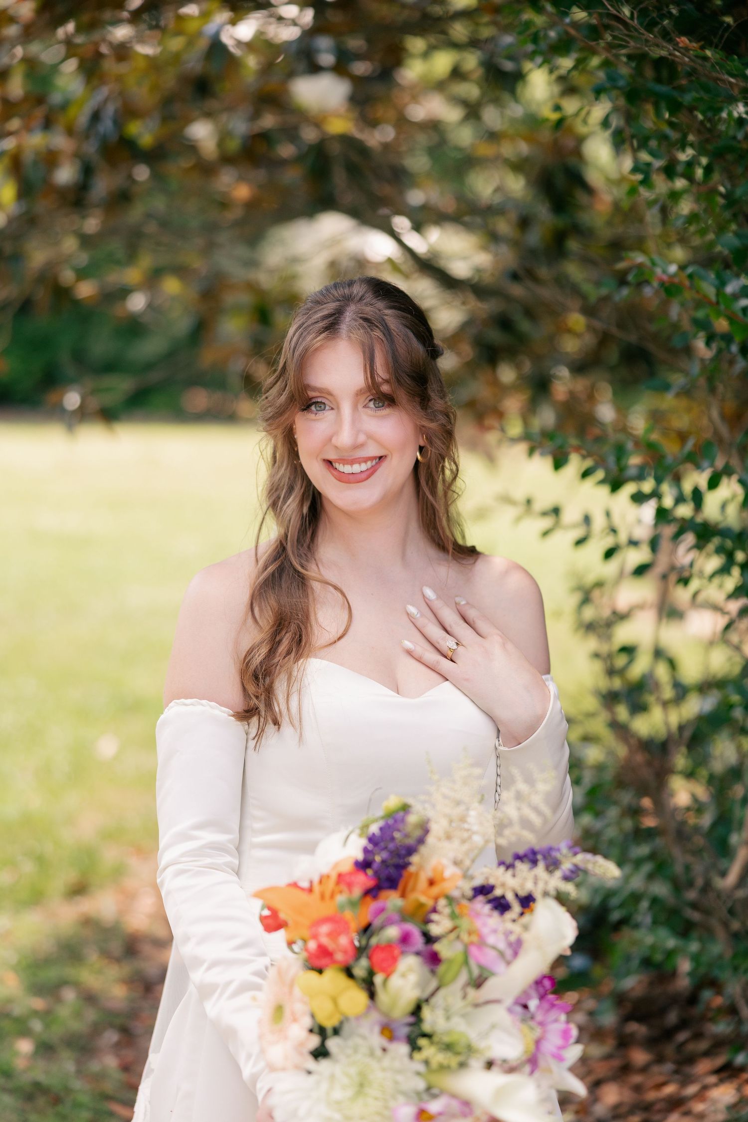 Smiling bride in off-shoulder white gown holds colorful bouquet while posing under lush green trees.