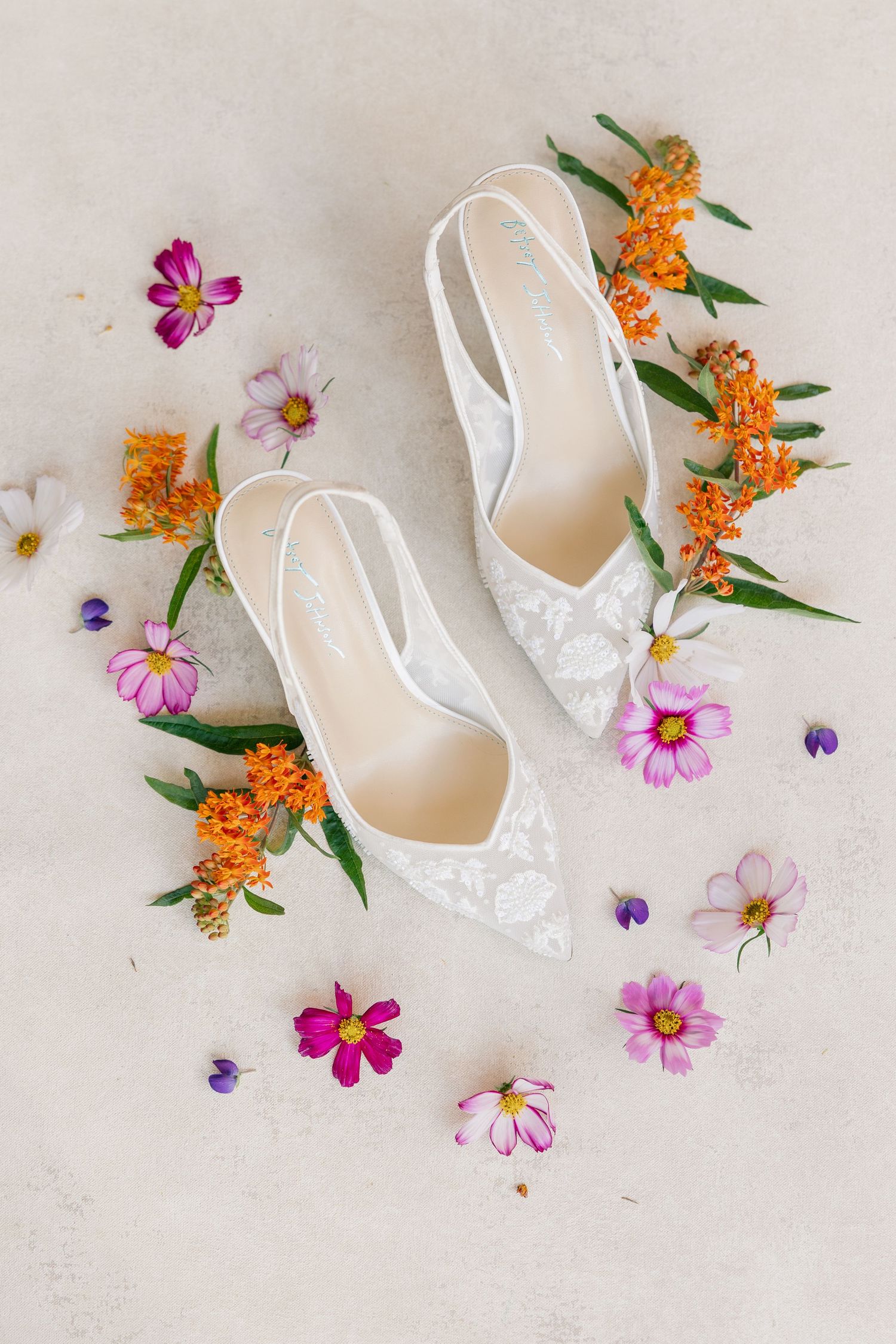 White lace bridal slingback heels surrounded by colorful pink and orange wildflowers on a light gray surface.