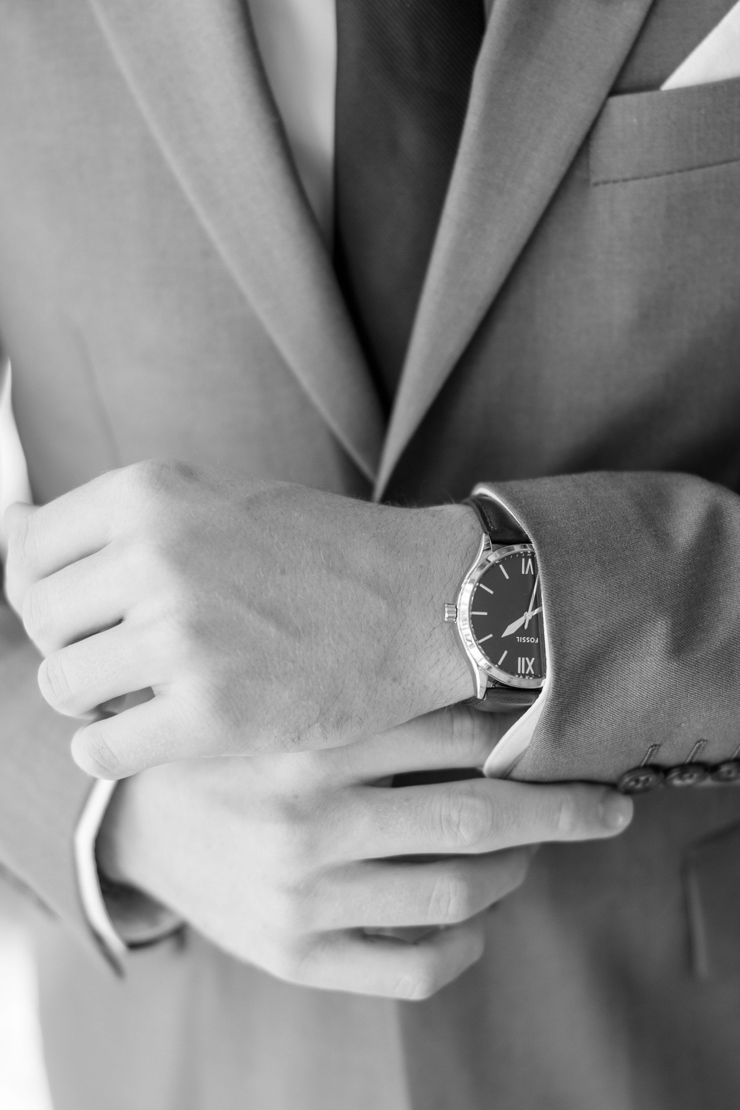 Close-up black and white photo of a groom adjusting his classic leather watch on his suit sleeve.