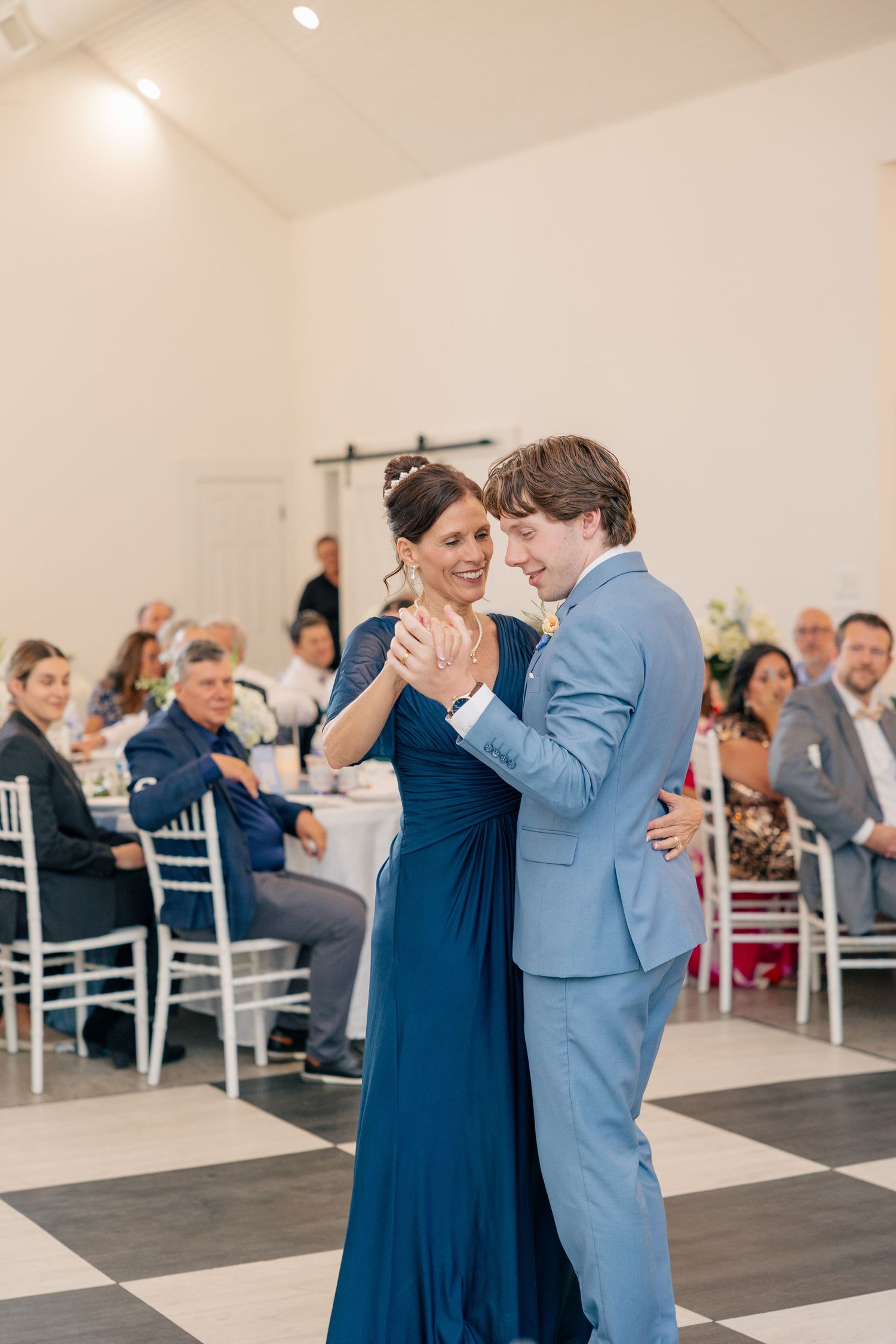 Groom in light blue suit shares mother-son dance at elegant wedding reception with checkered floor.