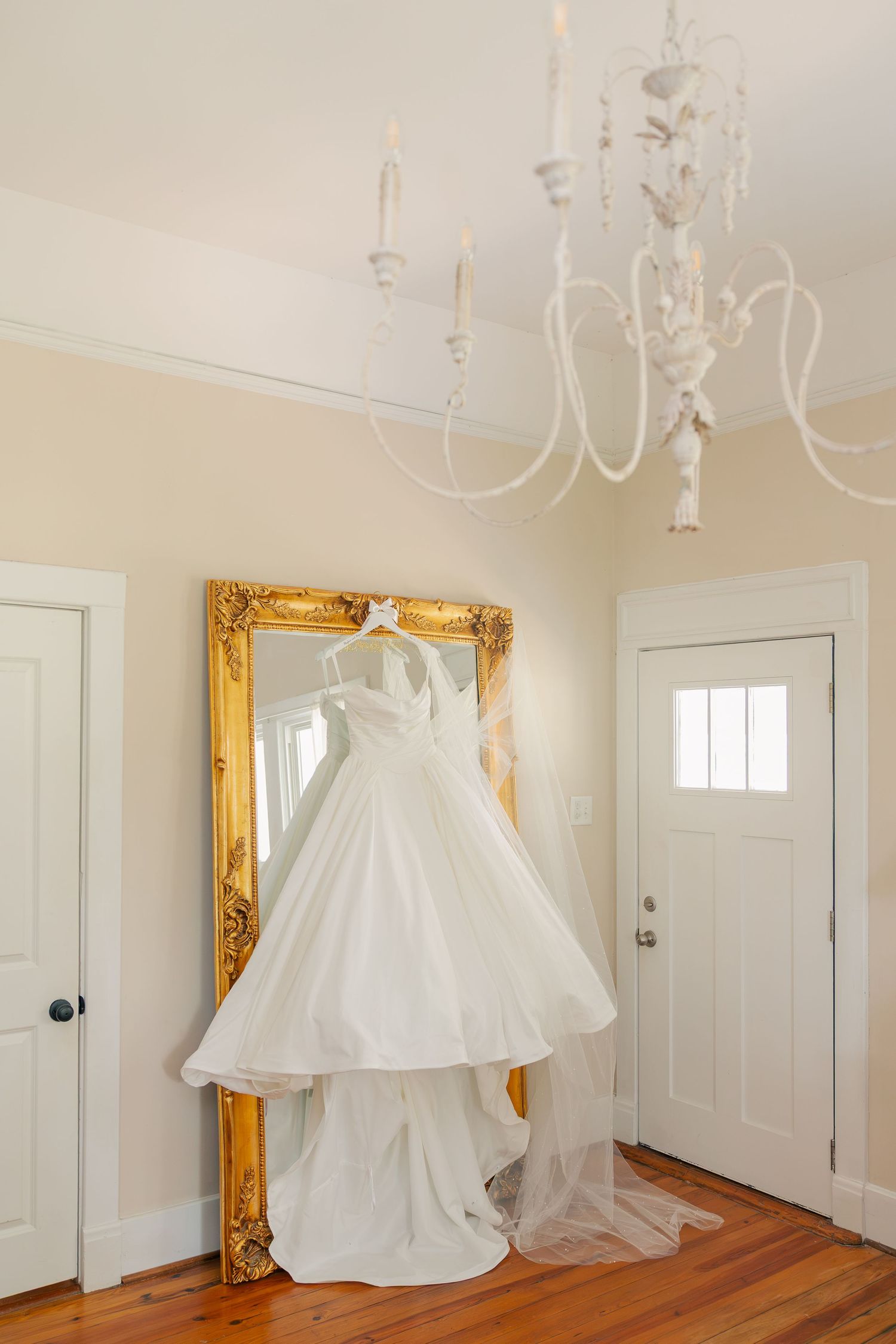 White wedding dress hanging on a gold ornate mirror in a bright entryway with white chandelier and hardwood floors.