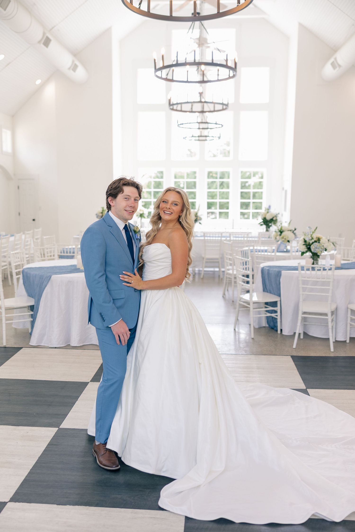 Bride and groom posing in elegant white reception hall with blue table linens and chandelier lighting.