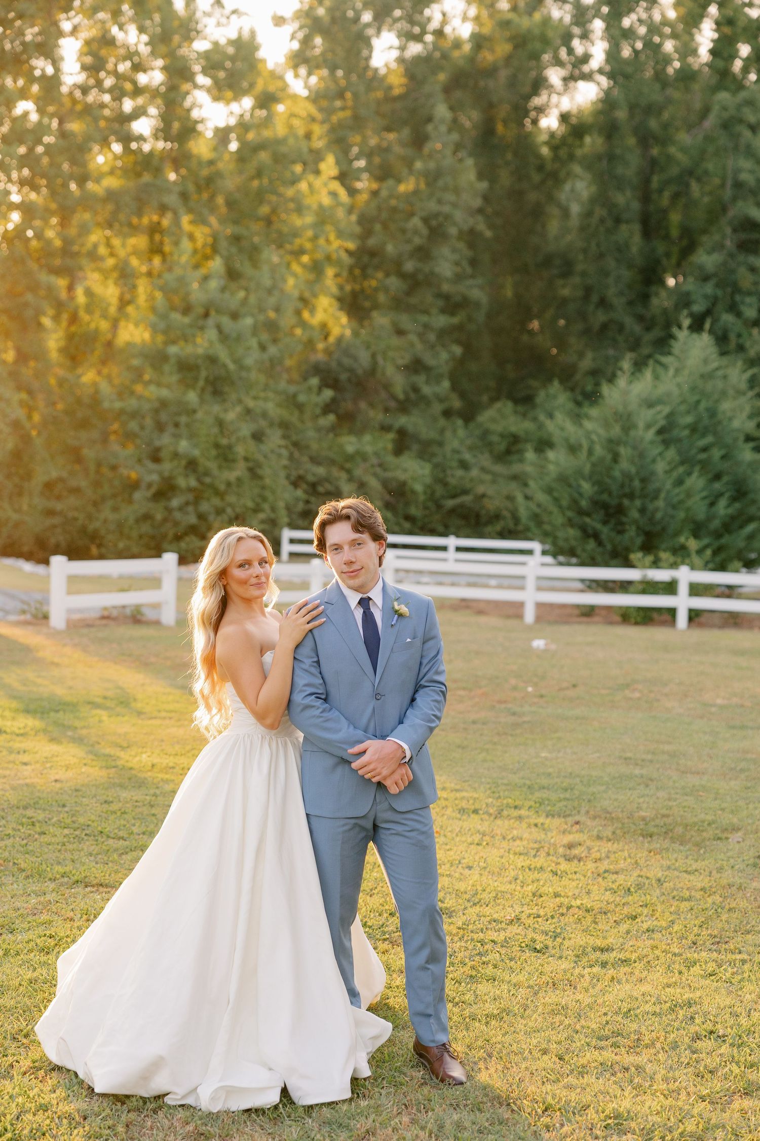 Bride in white gown and groom in light blue suit pose on green lawn at golden hour with white fence backdrop.