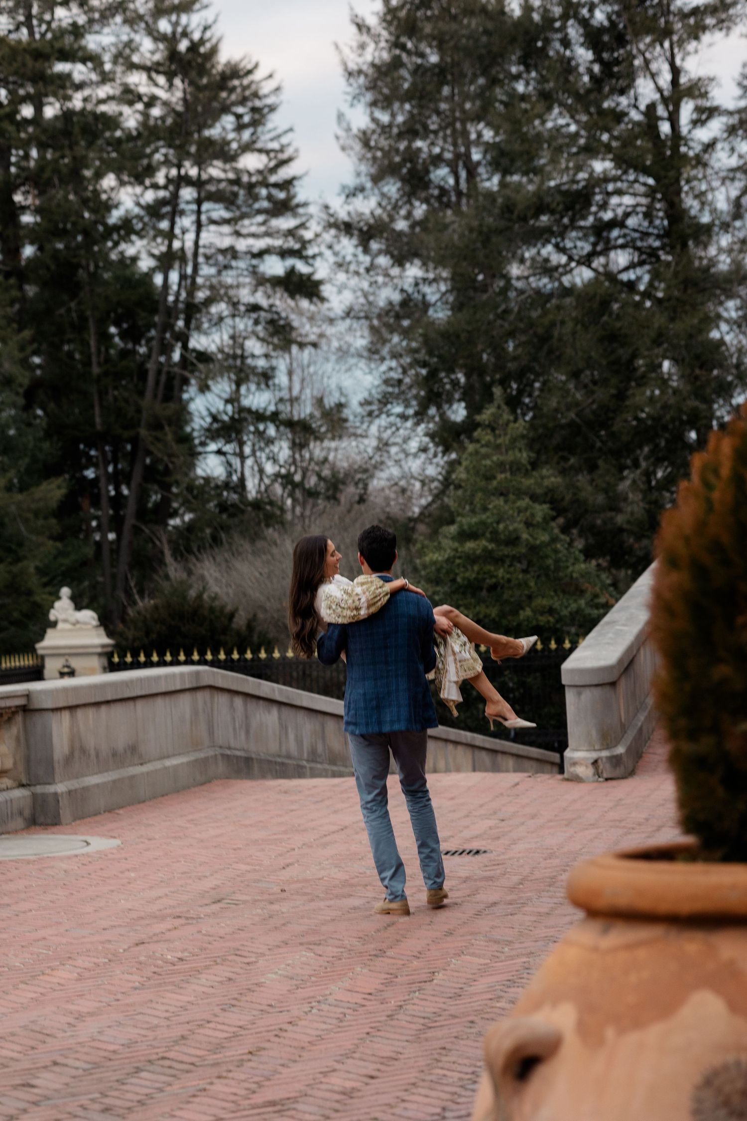 A couple enjoy a romantic twirling dance moment on a brick terrace surrounded by tall evergreen trees and stone railings.