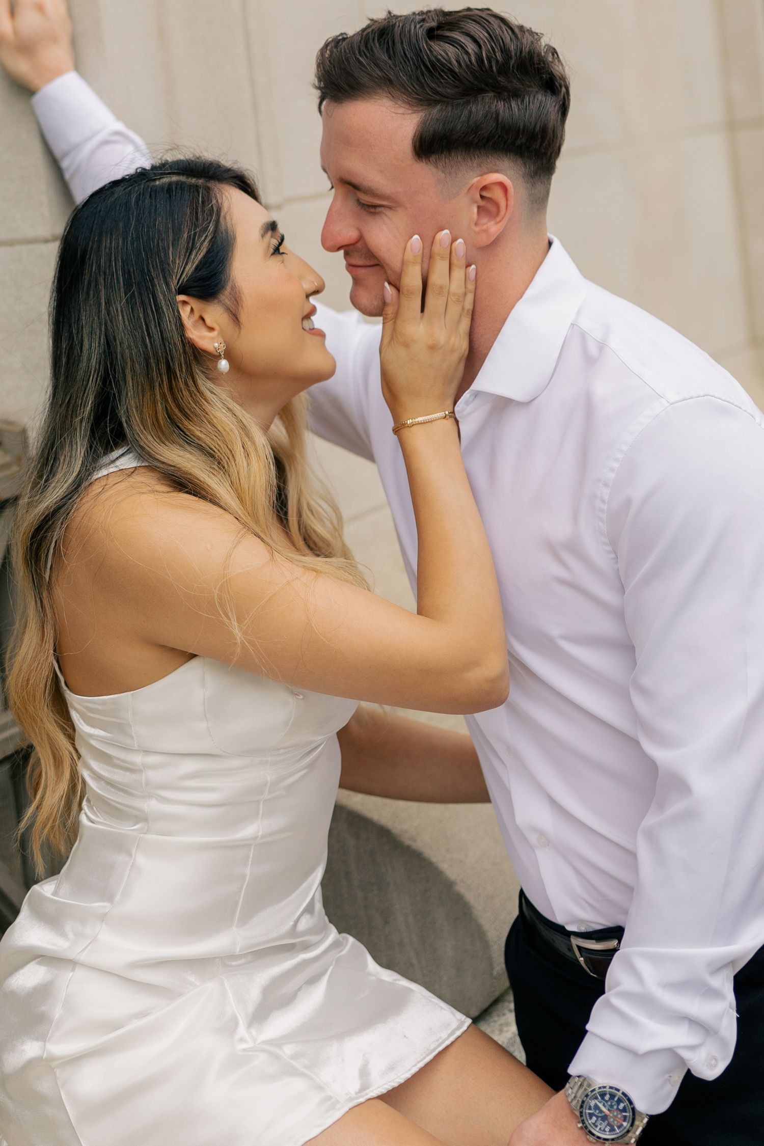 A couple in white attire shares intimate romantic moments in a series of candid poses against a neutral wall.