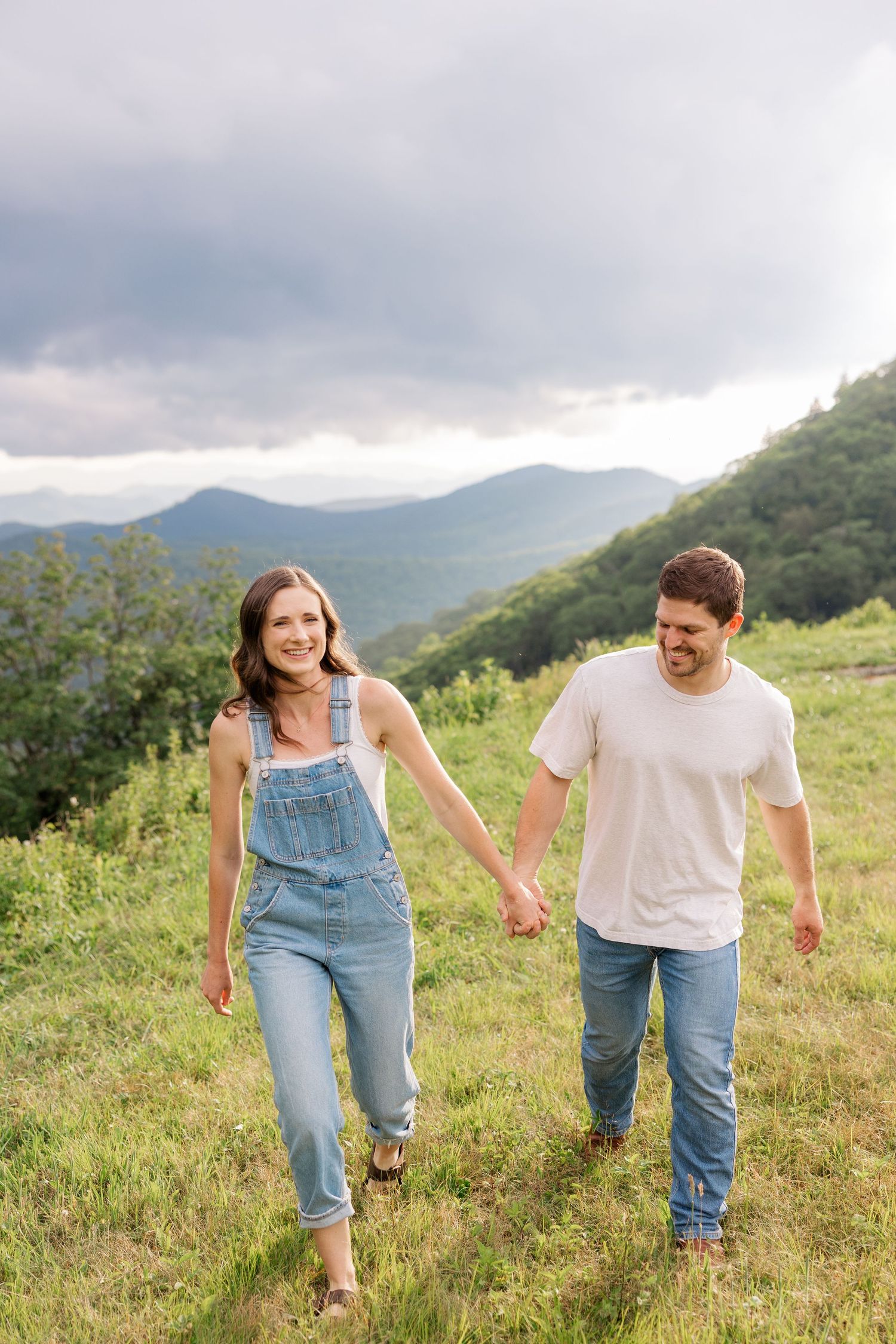 A couple runs playfully through a grassy mountain meadow wearing casual denim and white outfits.