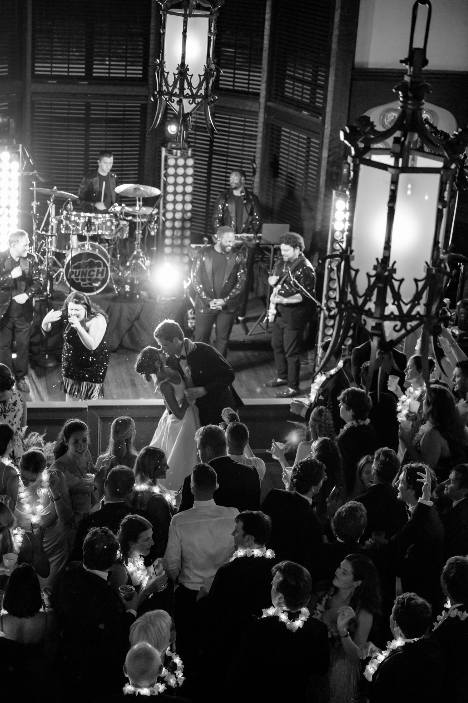 Black and white wedding reception photo with live band performing as couple shares first dance surrounded by guests.