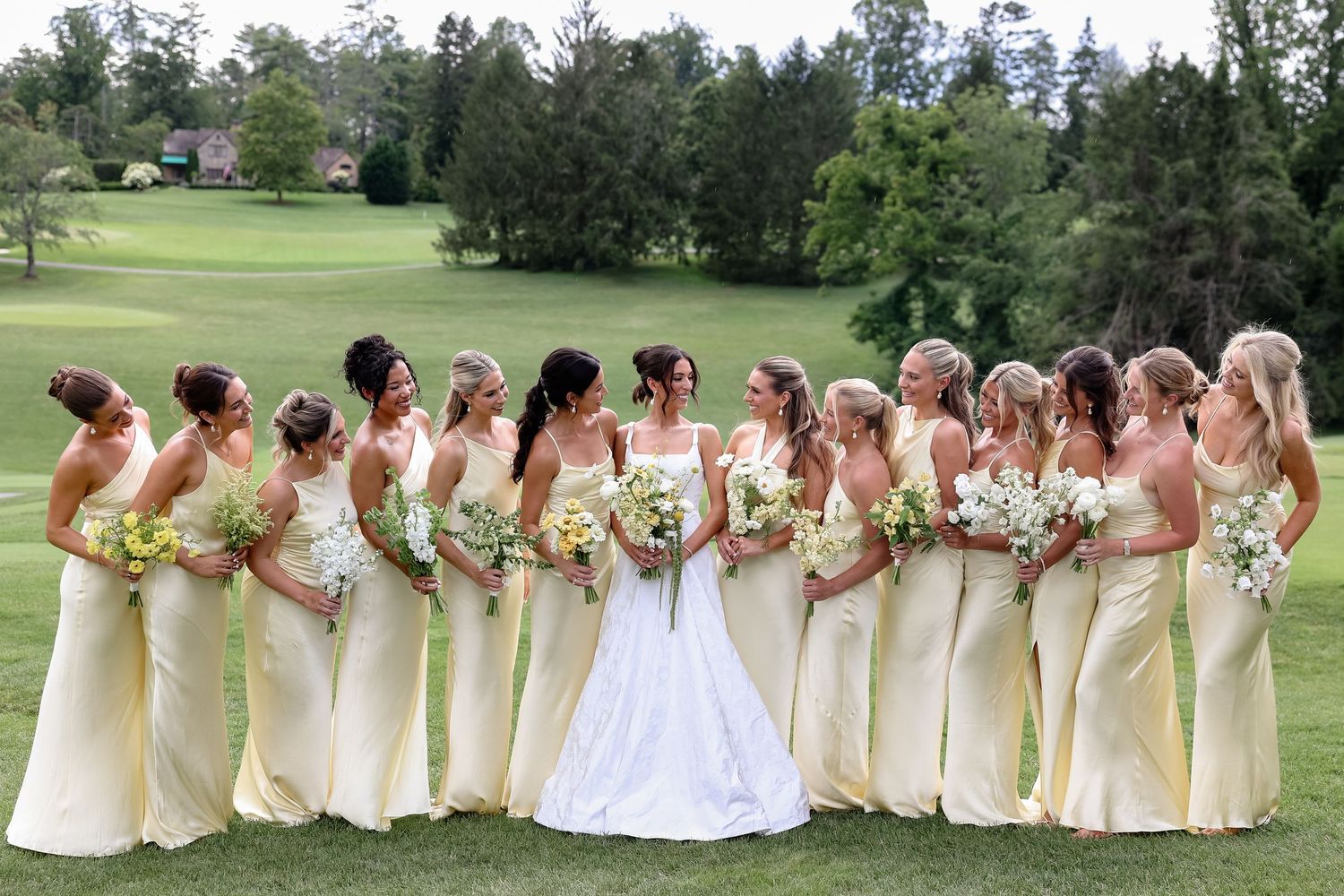 Bride in white gown stands with bridesmaids in yellow dresses holding bouquets on a green golf course lawn.