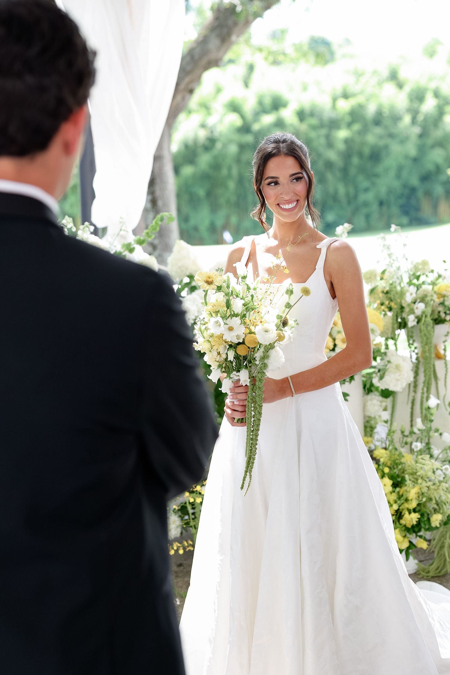 Smiling bride in sleeveless white gown holds yellow and white bouquet at outdoor wedding ceremony.