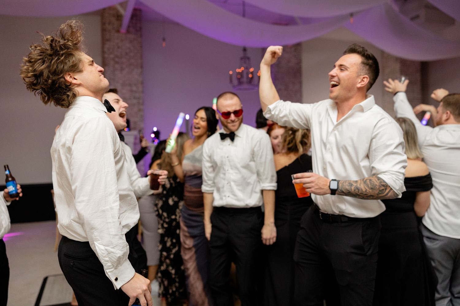 Wedding guests dancing enthusiastically at a reception with purple lighting and draped white fabric decor.