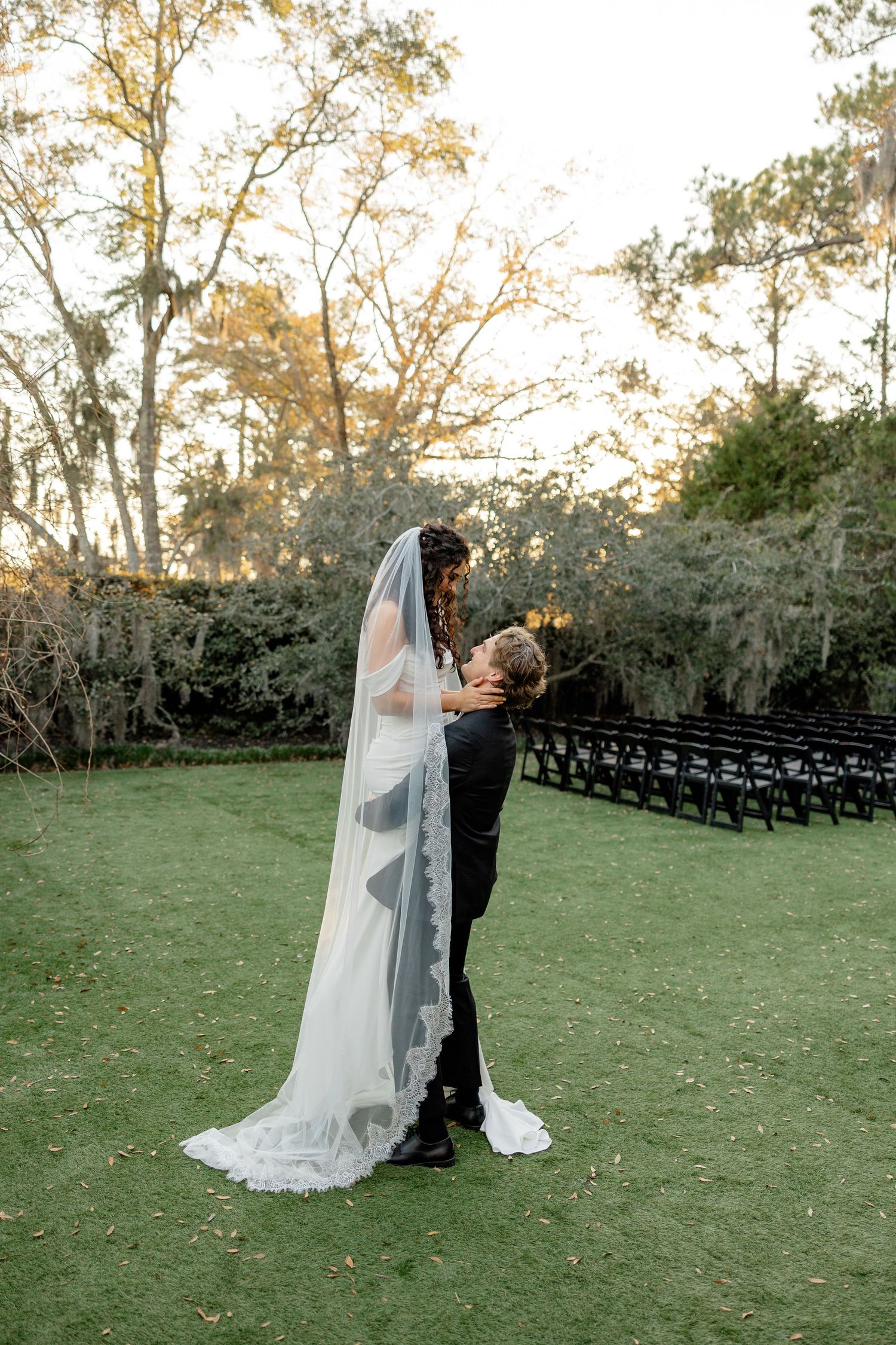 A couple embraces during their wedding photo session on a lawn with ethereal backlit veil.
