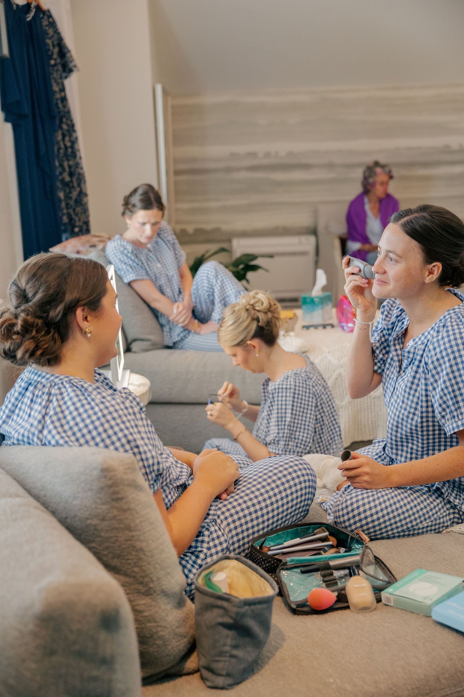 Bridesmaids in blue gingham pajamas applying makeup together on couch before wedding.