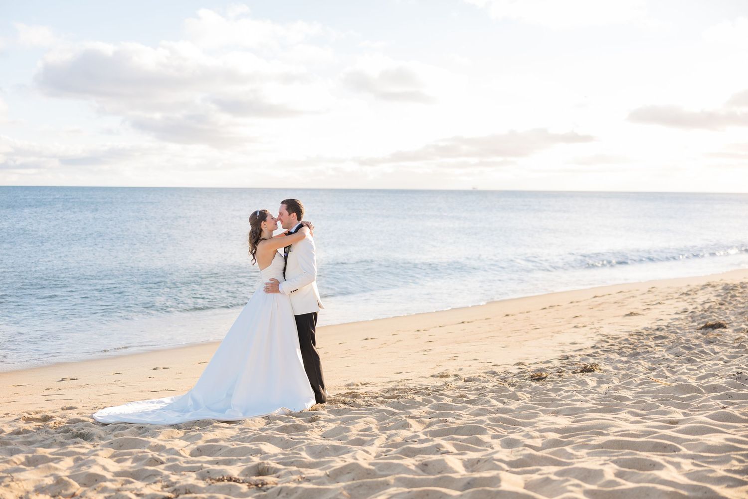 Bride and groom sharing a kiss on a sandy beach with calm blue ocean at sunset.