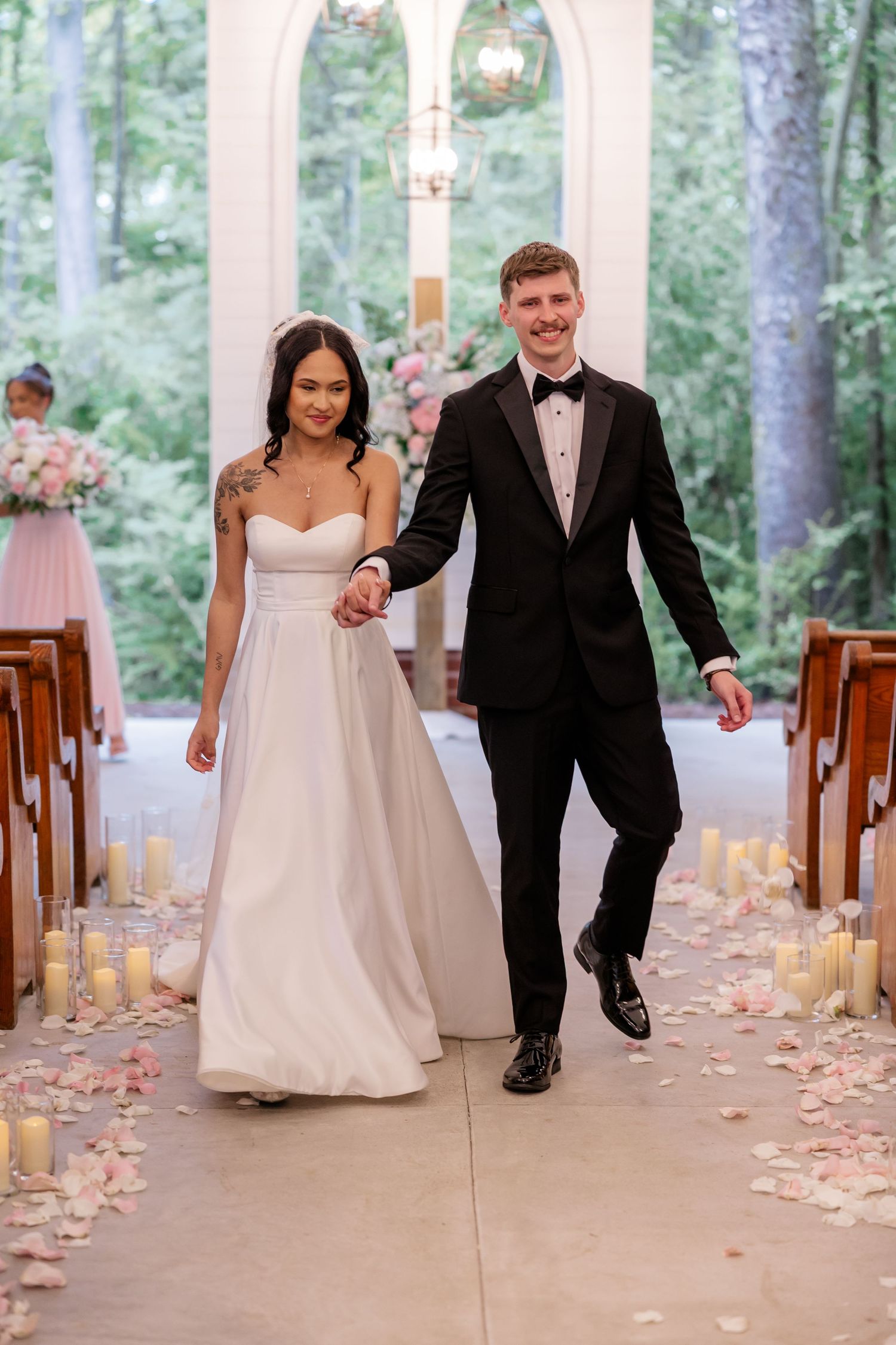 Newlyweds walking down candlelit aisle in elegant chapel surrounded by flower petals.