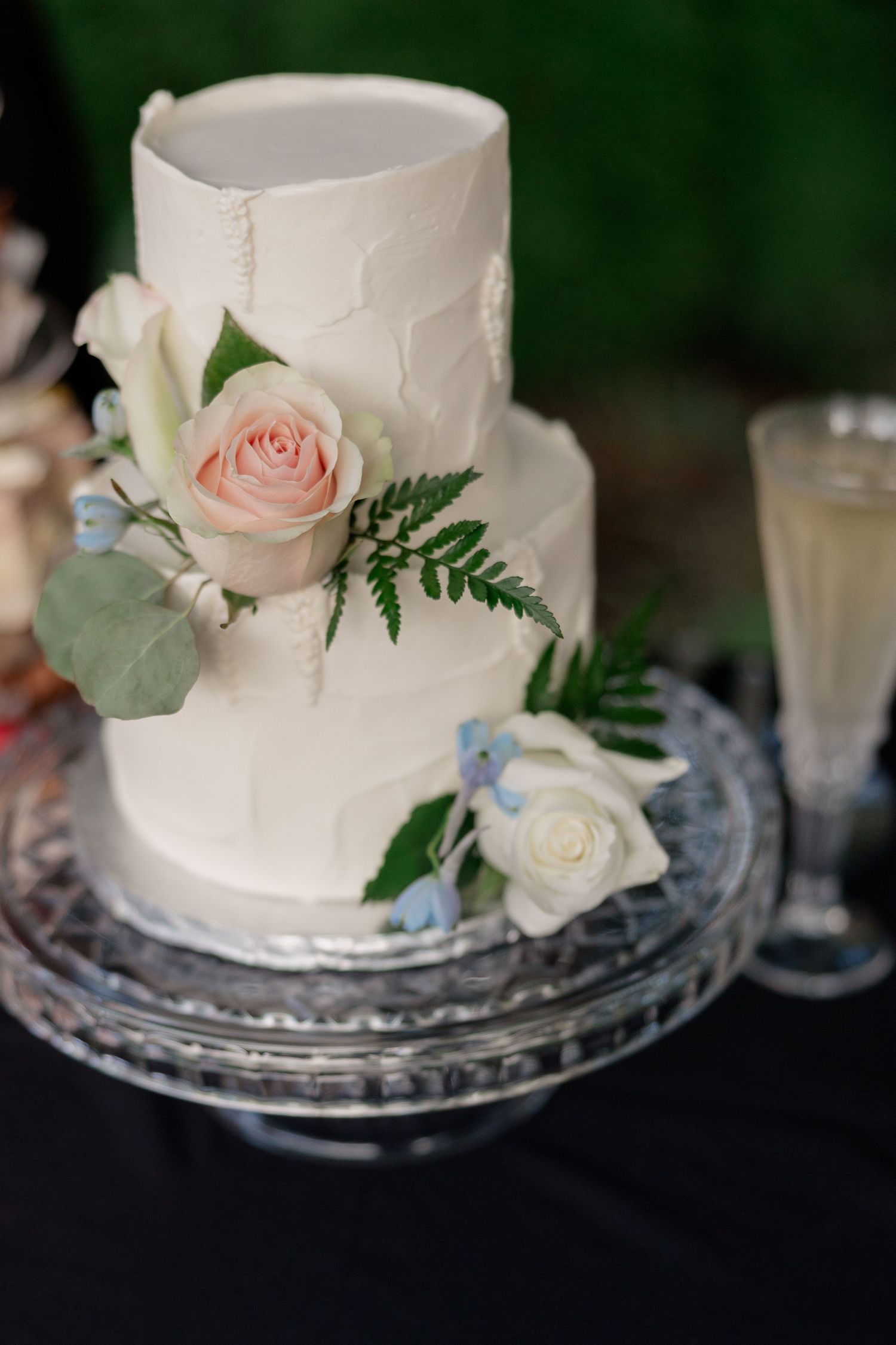 Simple white wedding cake decorated with blush roses, ferns, and delicate floral accents on a glass stand.