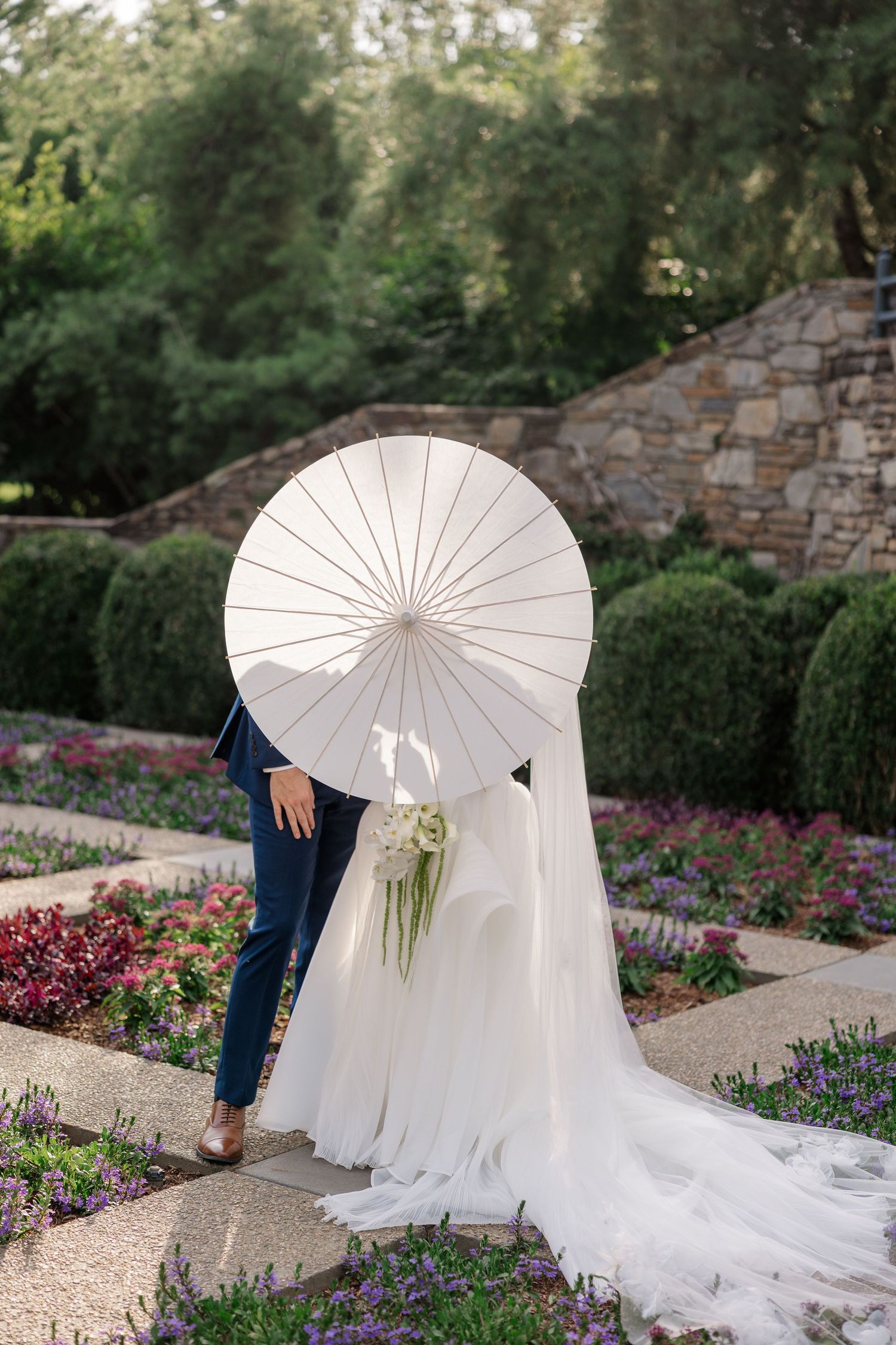 A white parasol frames an elegant wedding dress train in a manicured garden.