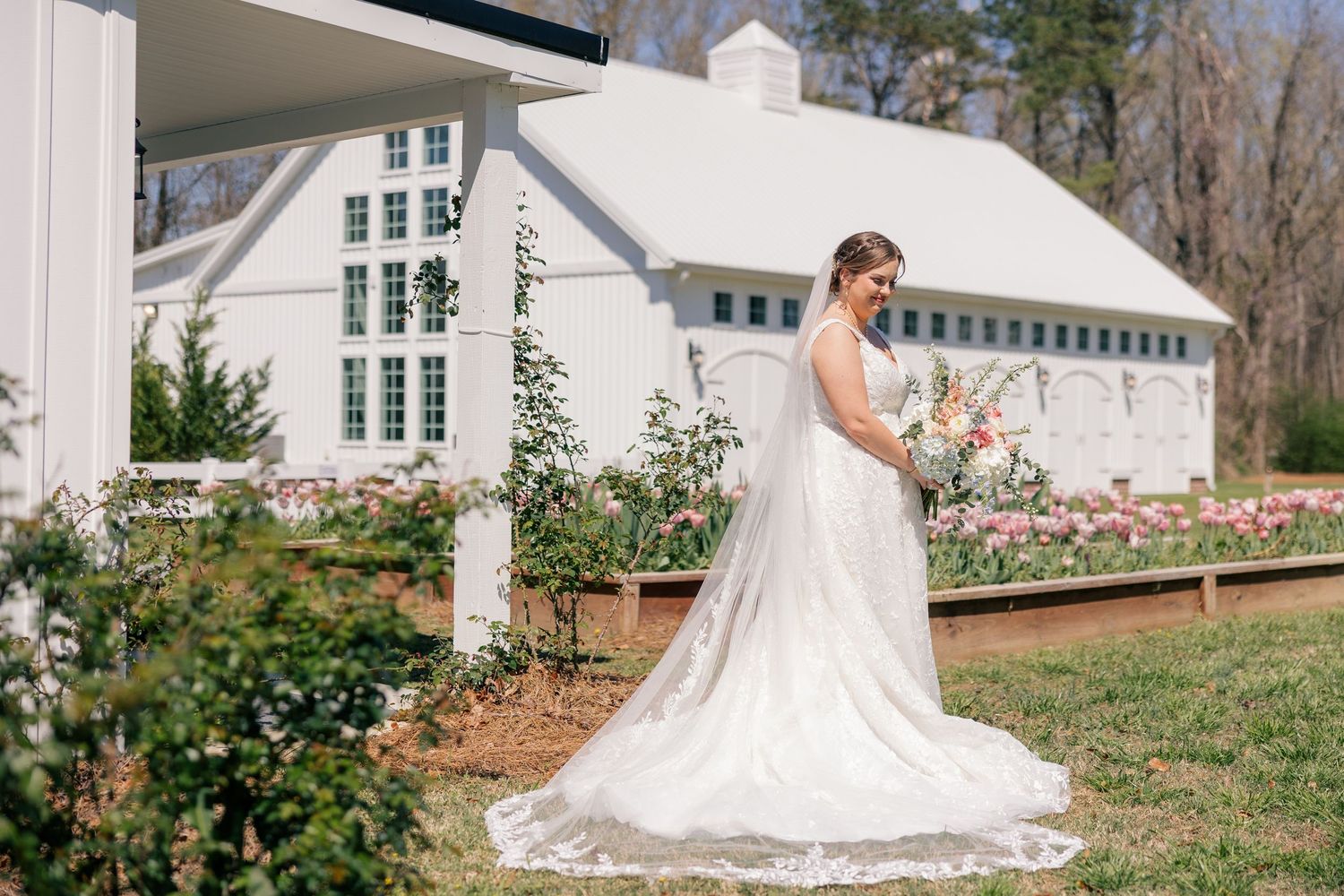 Elegant bride in white wedding gown poses gracefully in front of a white building on a sunny day.