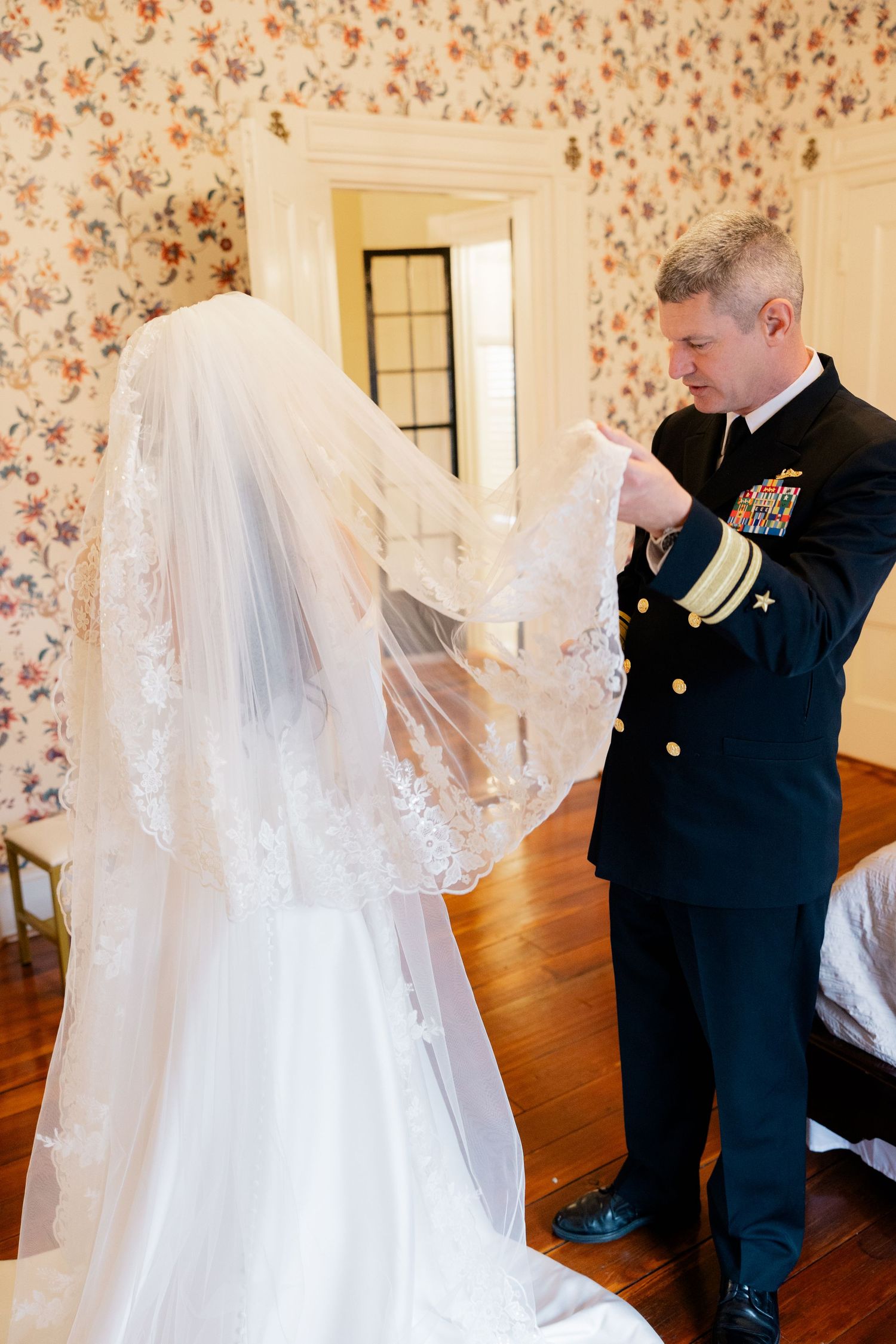 Wedding preparation scene showing intricate details of bridal veil and dress against floral wallpaper backdrop.
