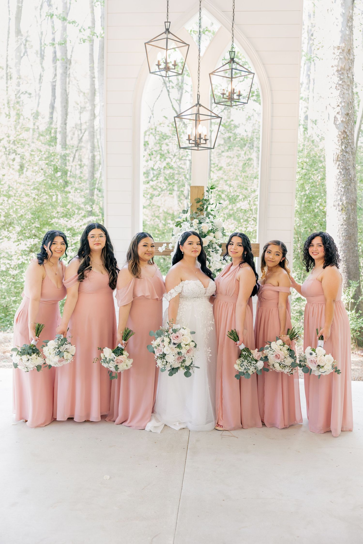 Bridesmaids in matching pink dresses holding white floral bouquets pose in an elegant white venue.