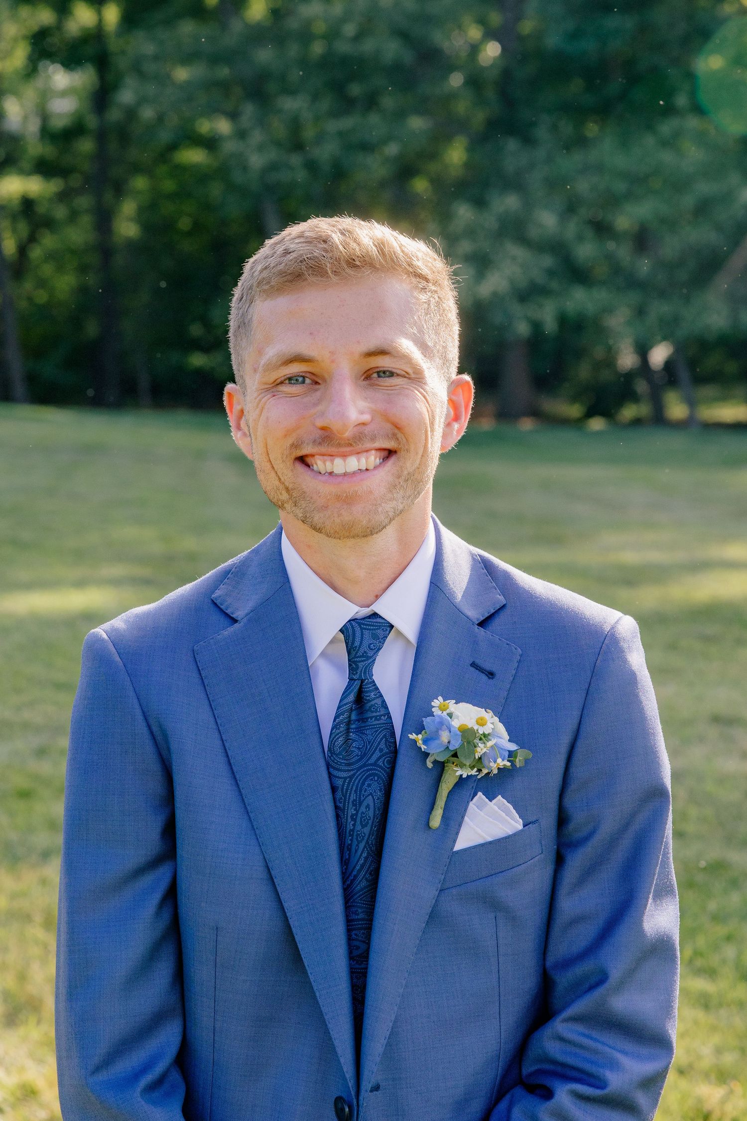 Smiling groom in blue suit with floral boutonniere poses outdoors on green lawn.