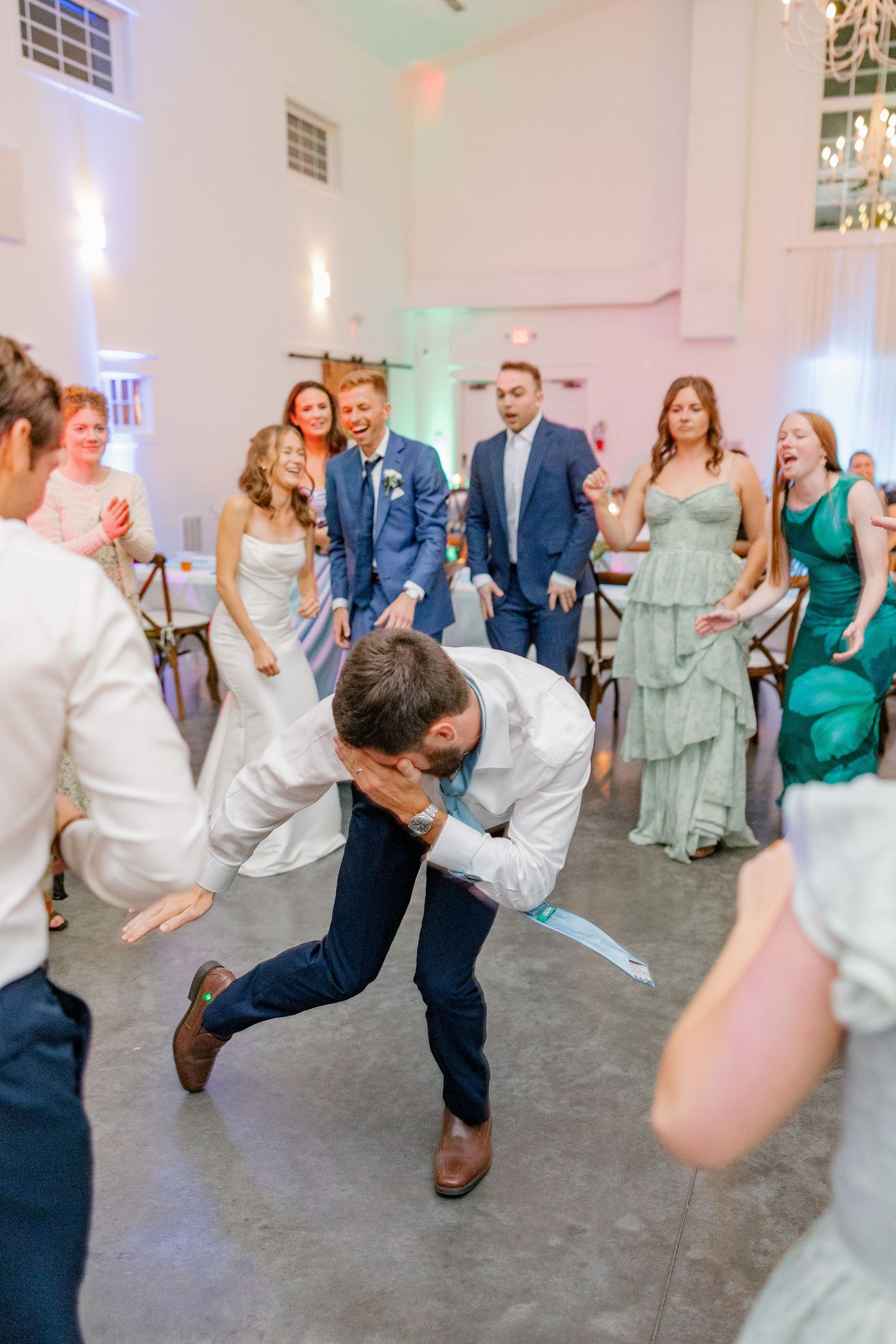 Wedding guests cheer as a man energetically dances at a lively indoor wedding reception.