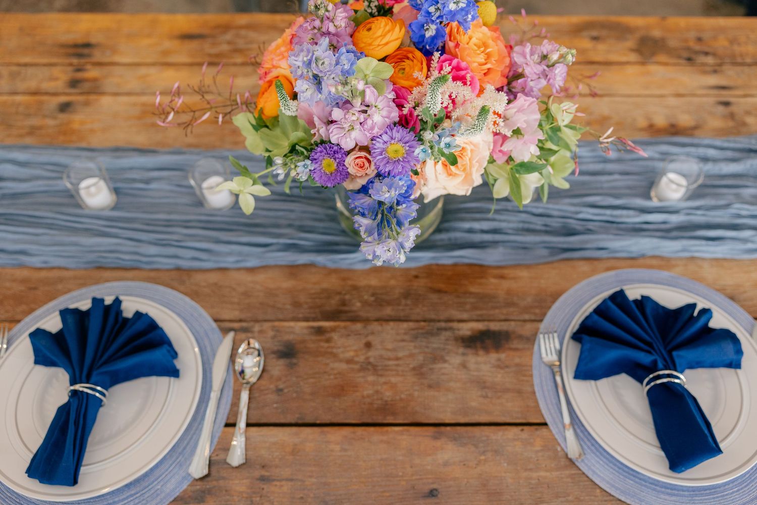 Rustic wooden reception table set with blue napkins and a colorful wildflower centerpiece arrangement.