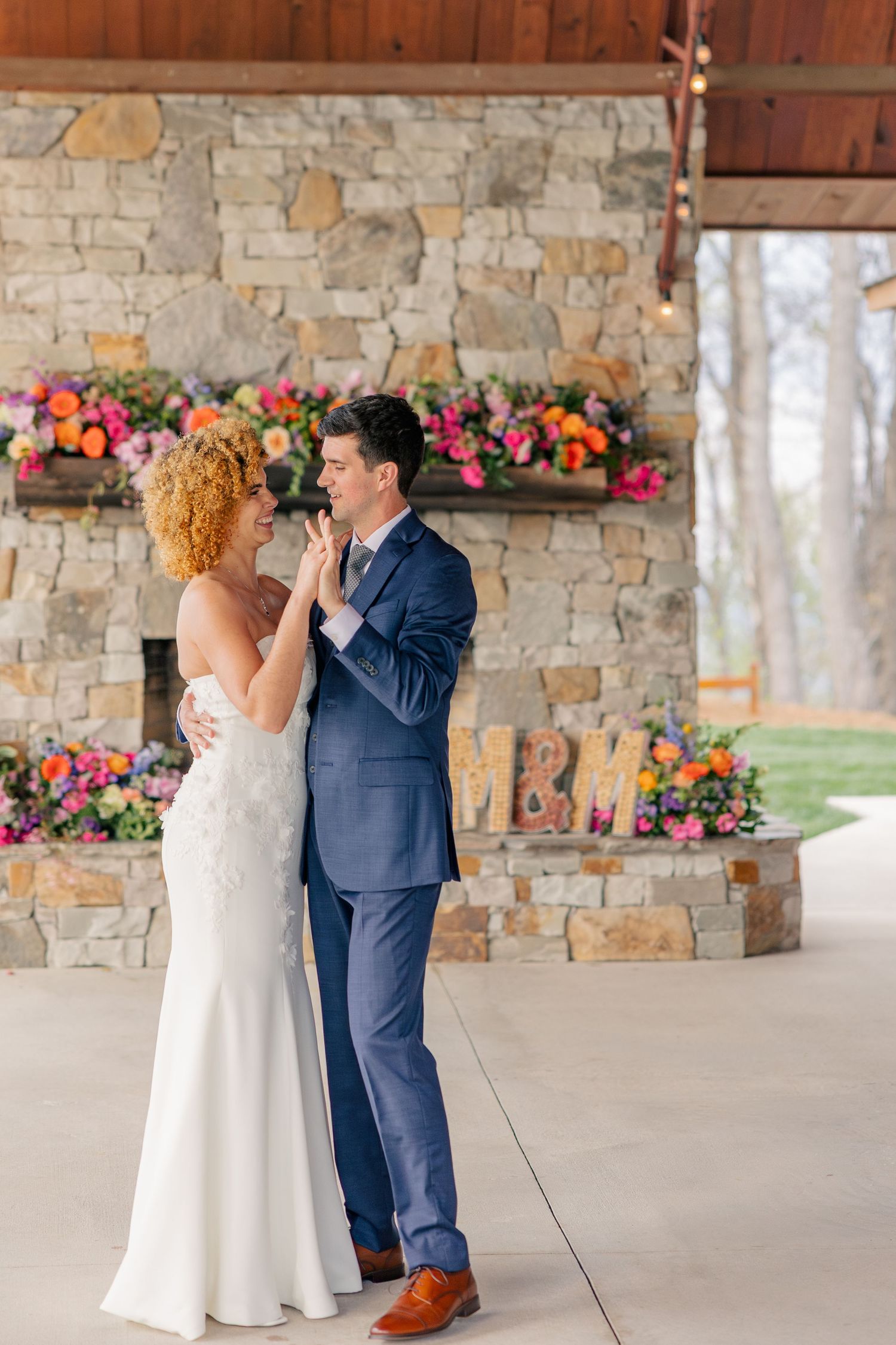 Bride and groom share first dance under a covered pavilion with stone fireplace and colorful floral decorations.