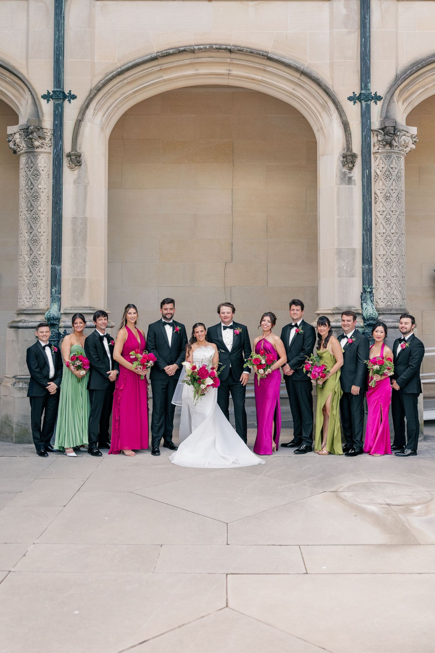 Wedding party in pink and green gowns with black tuxedos posed under stone archway.