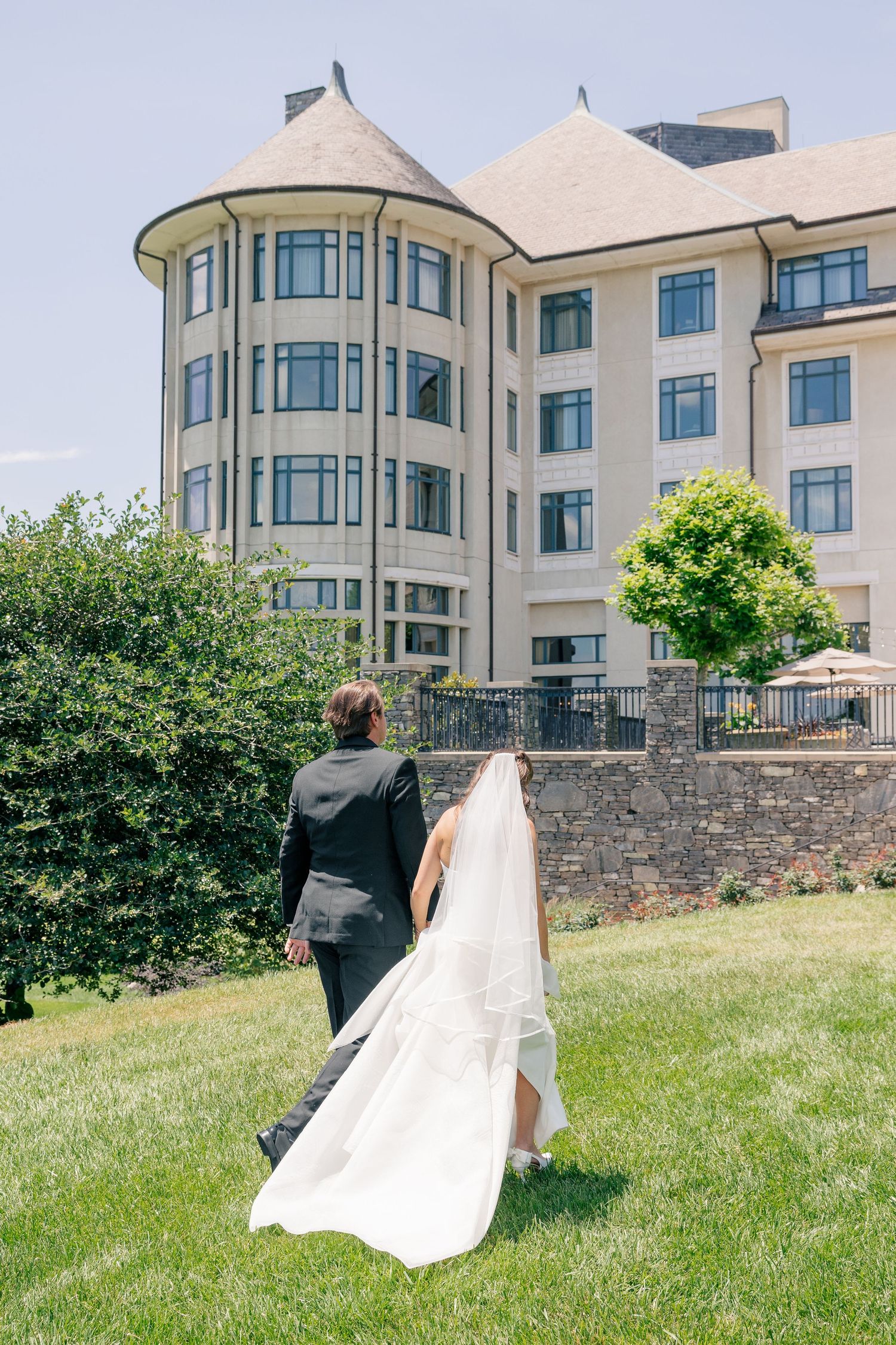 Bride and groom walk hand in hand across green lawn toward elegant multi-story wedding venue.
