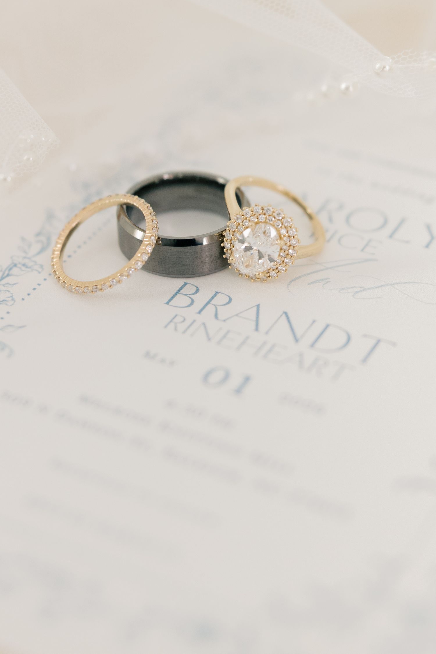 Three wedding rings on invitation card for Brandt Rineheart, featuring diamond halo engagement ring and black band.