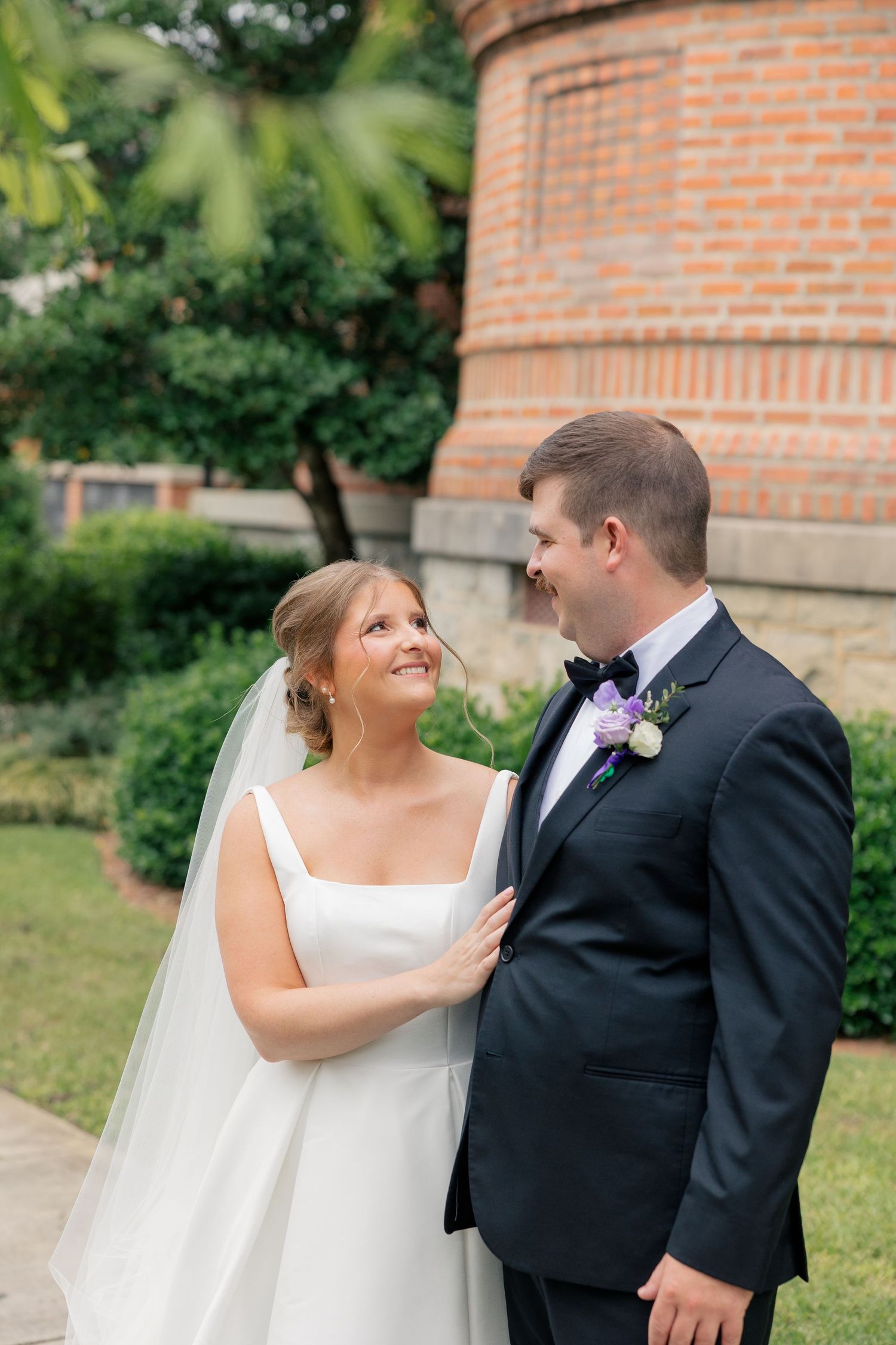 Smiling bride in white gown with veil gazes at groom in navy suit outdoors.