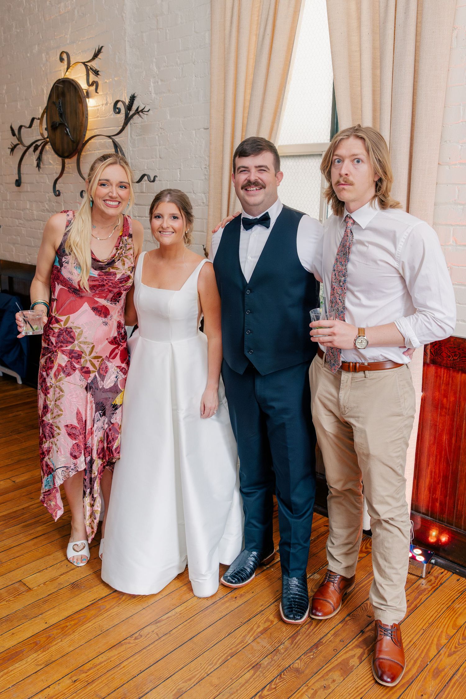 Bride in white gown and groom in navy vest pose with two guests at an elegant indoor wedding reception.