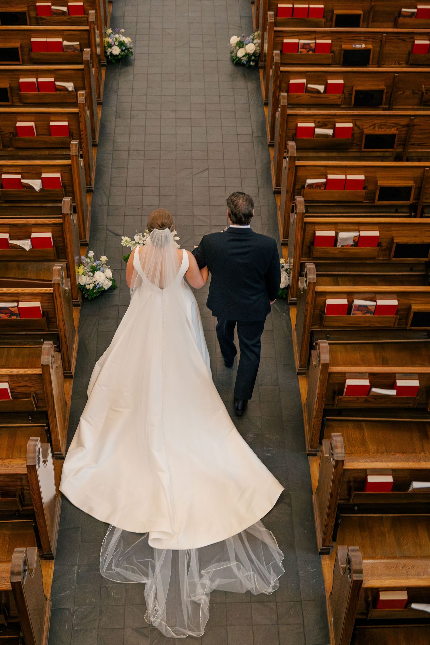 Aerial view of bride in elegant white gown walking down church aisle with escort between wooden pews.