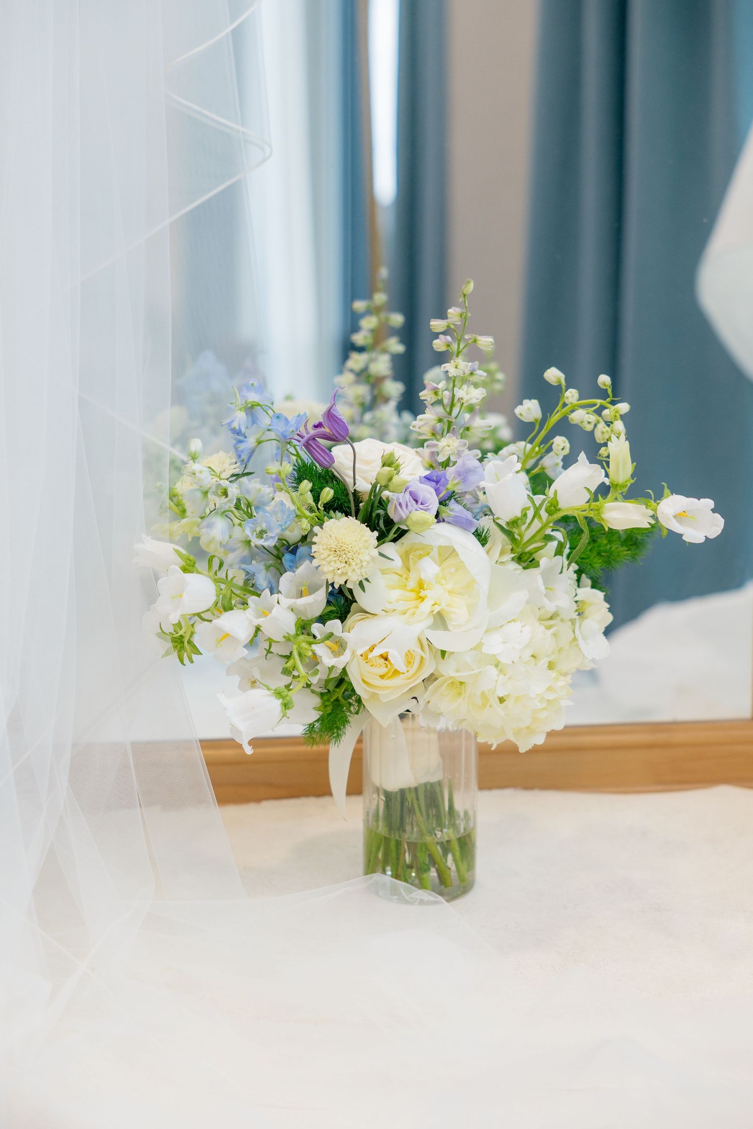 Elegant wedding bouquet with white roses and purple flowers in a glass vase near a window with sheer curtains.