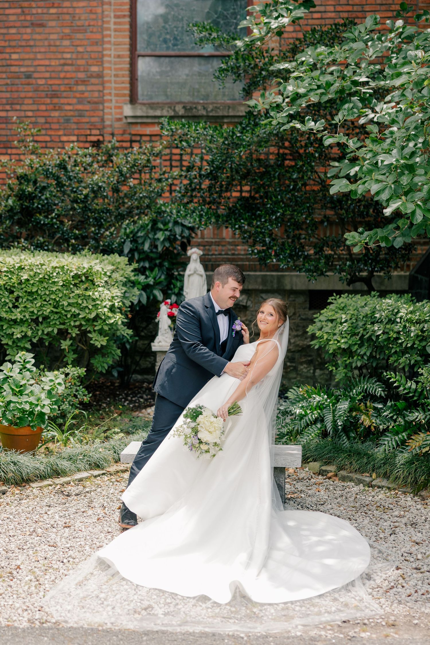 Bride and groom share romantic embrace in lush garden beside brick church with white statue backdrop.