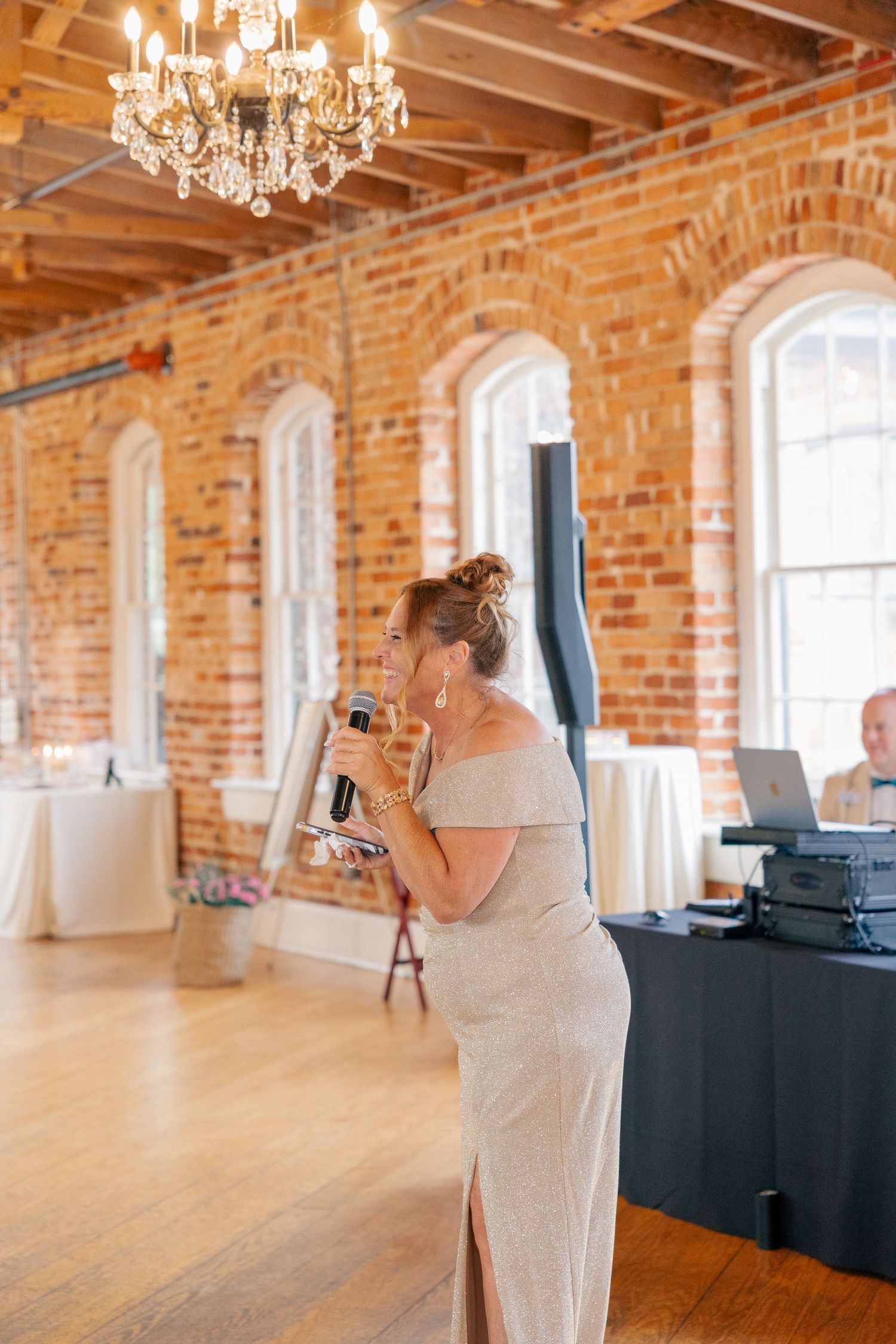 A woman in an off-shoulder gown gives a speech holding a microphone in an elegant brick-walled wedding venue.