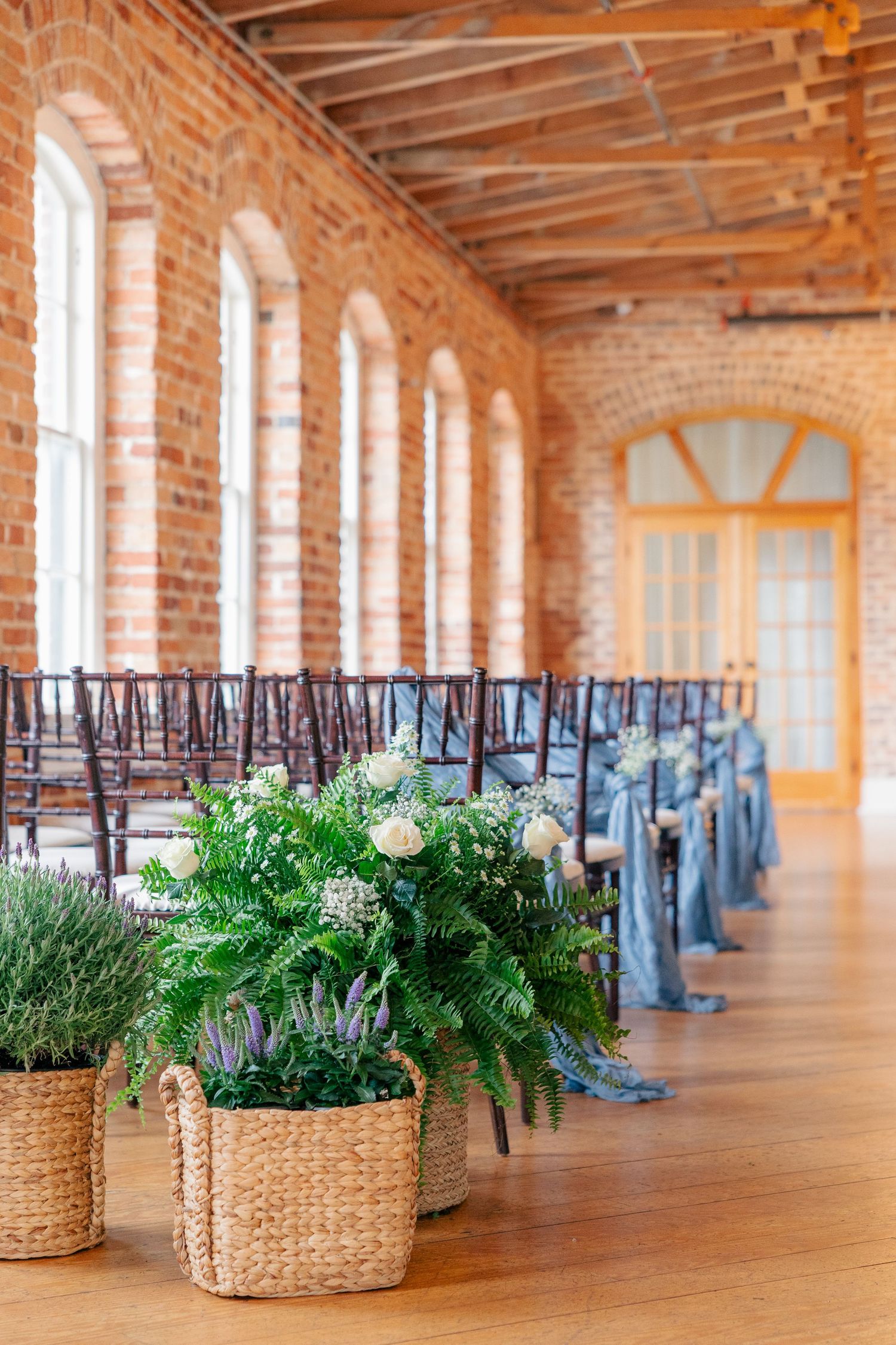 Elegant wedding ceremony venue with brick walls, wood floors, floral baskets, and blue ribbon chairs.