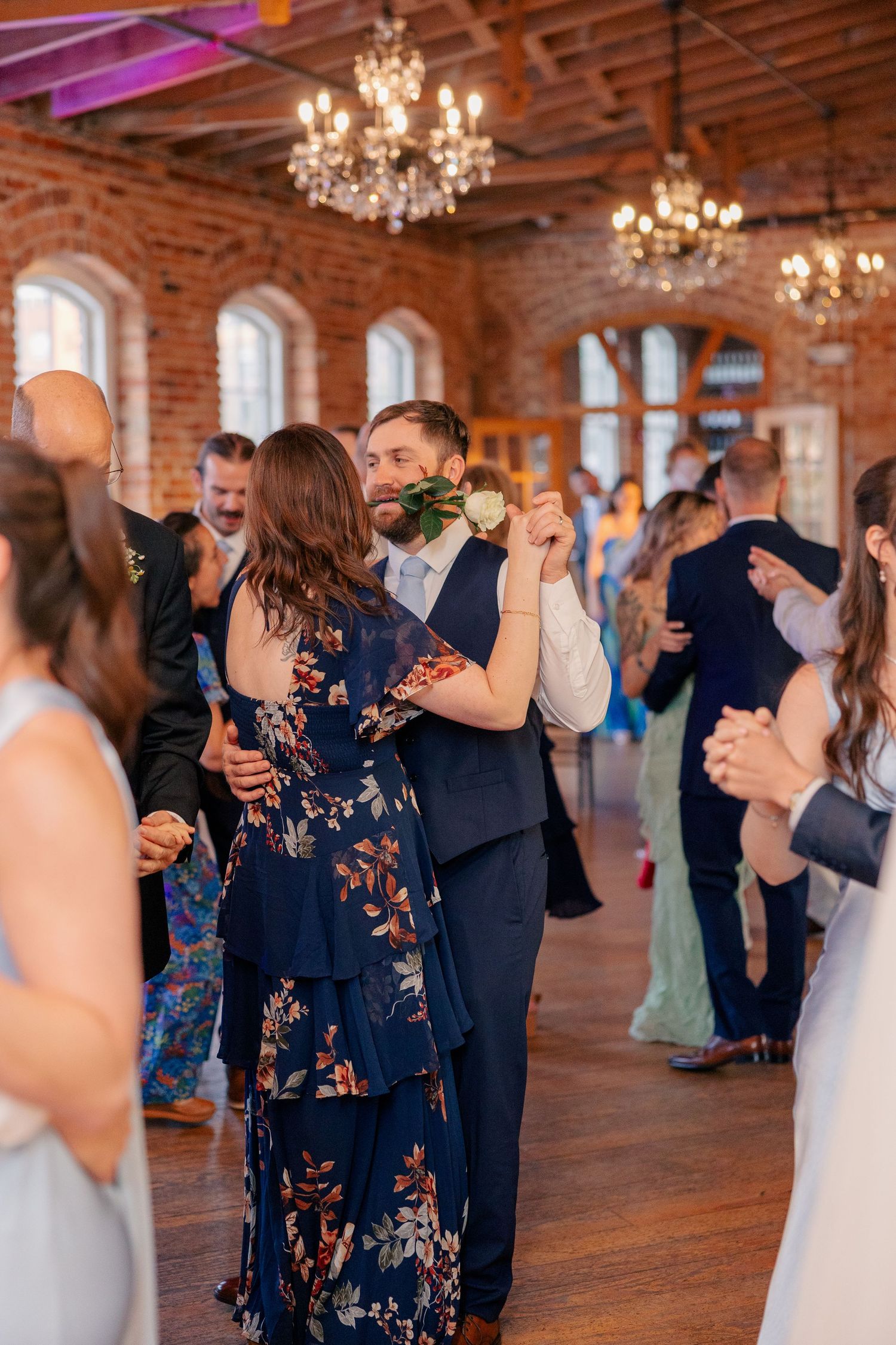 Wedding guests dance joyfully in a warmly lit rustic brick ballroom with crystal chandeliers above.