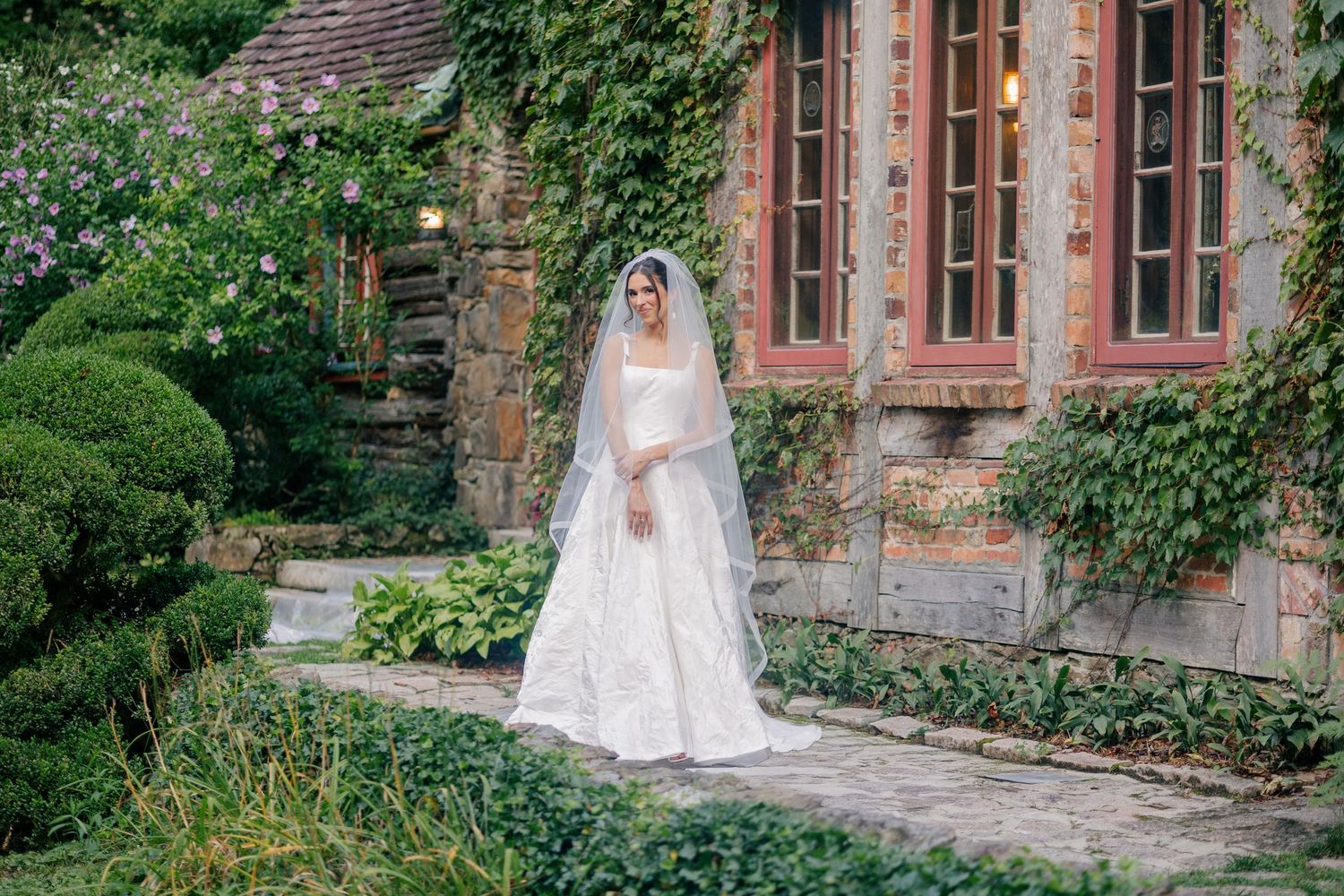 A figure in a beautiful white wedding dress poses in a garden setting next to a historic stone building with ivy-covered walls.