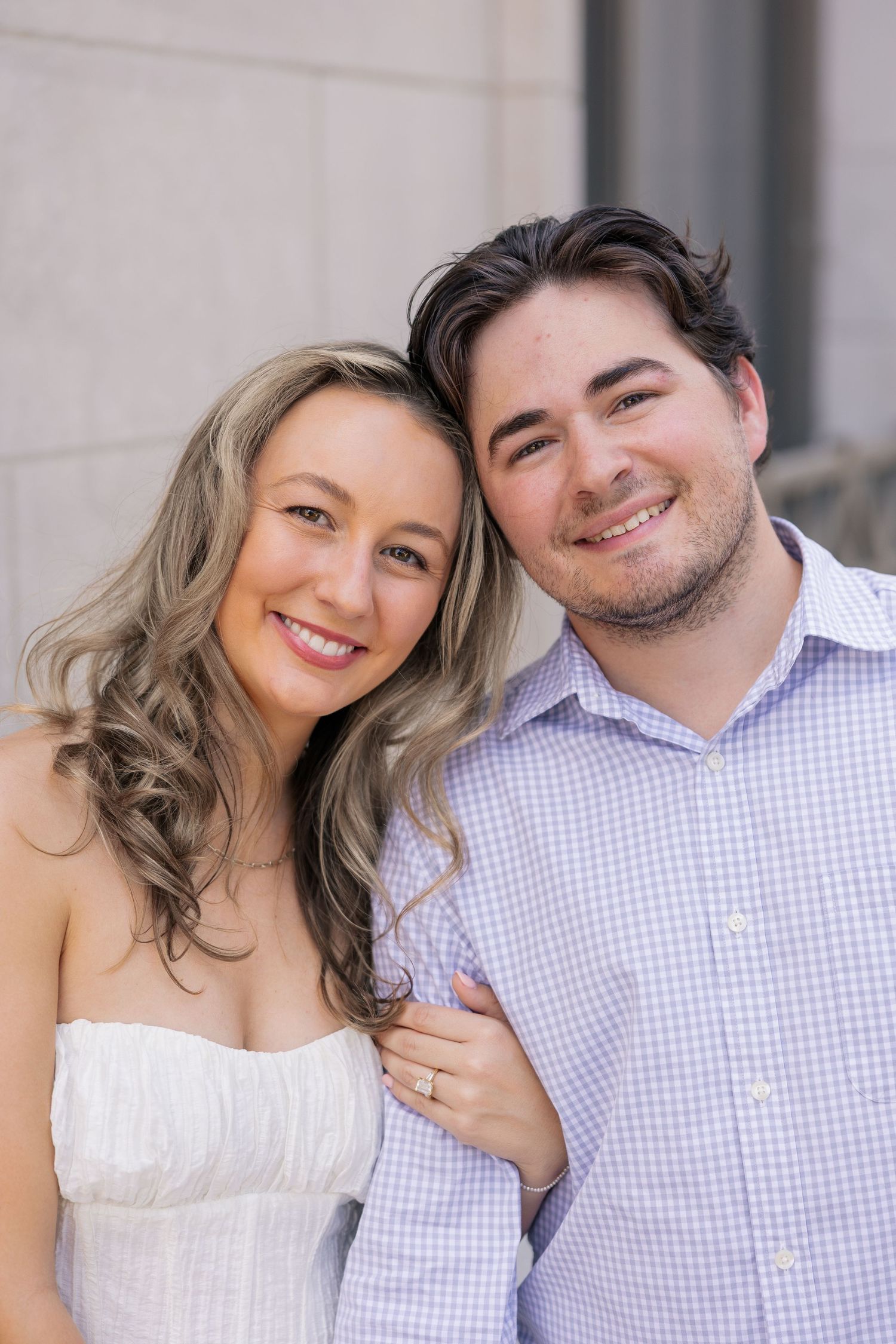 A smiling couple poses closely together, woman in white dress with curly hair, man in light blue checkered shirt.