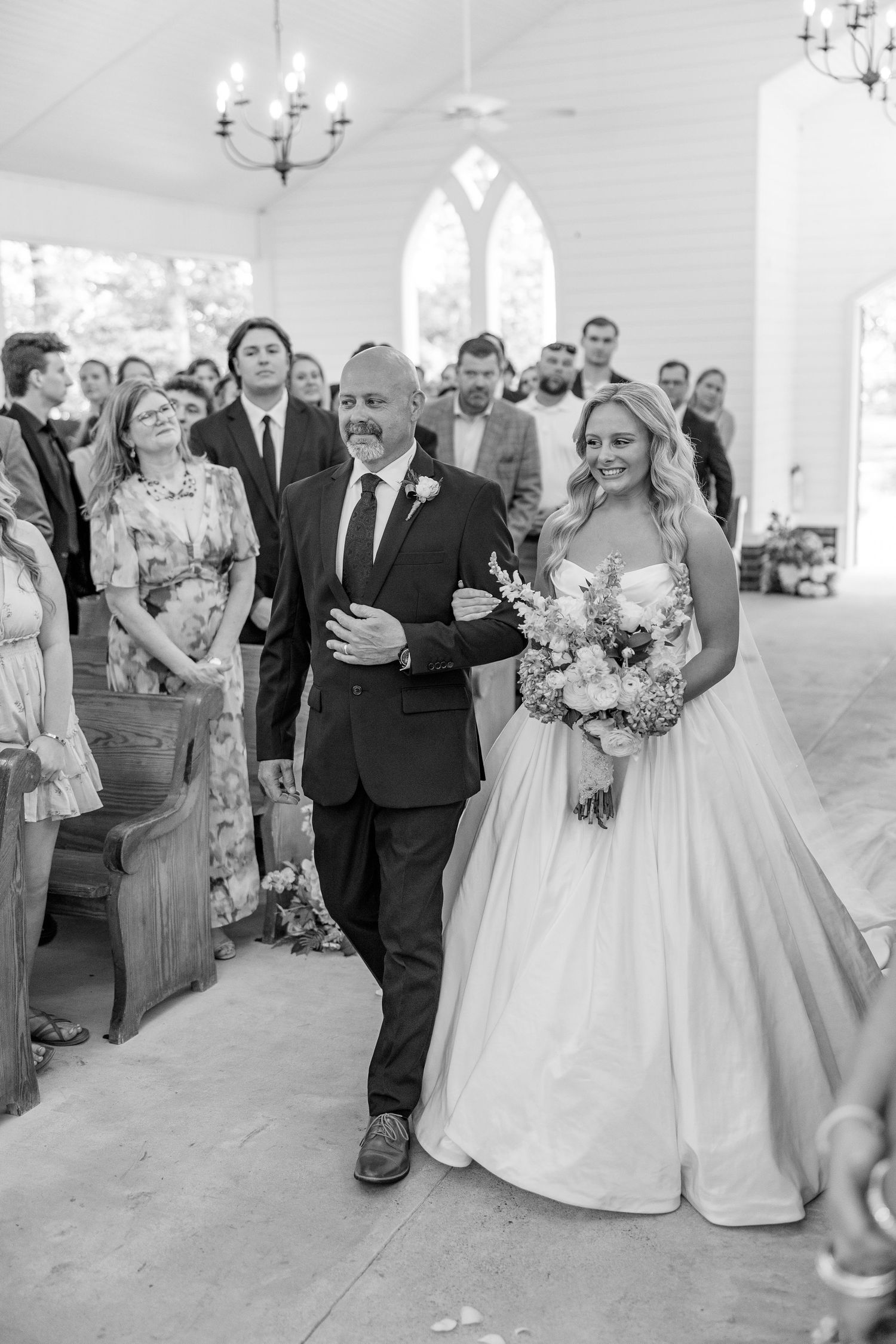 Black and white photo of smiling bride walking down chapel aisle with her father.
