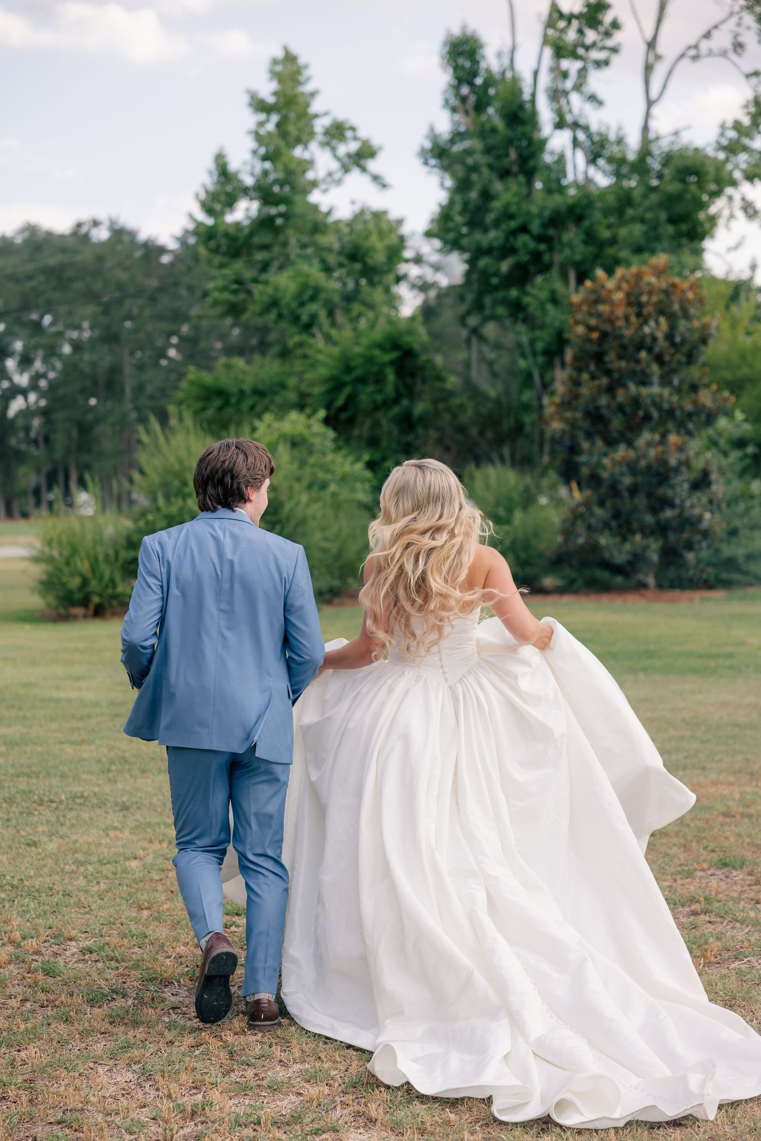 Bride and groom walking hand in hand through lush green garden, bride in flowing white gown, groom in light blue suit.
