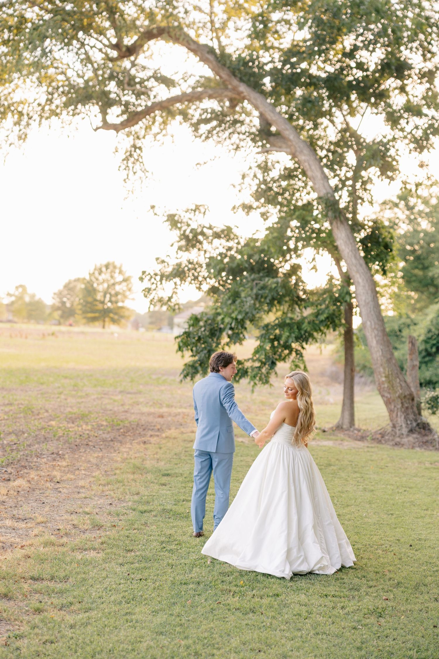 Bride and groom hold hands walking through sunlit garden with large oak trees at golden hour.