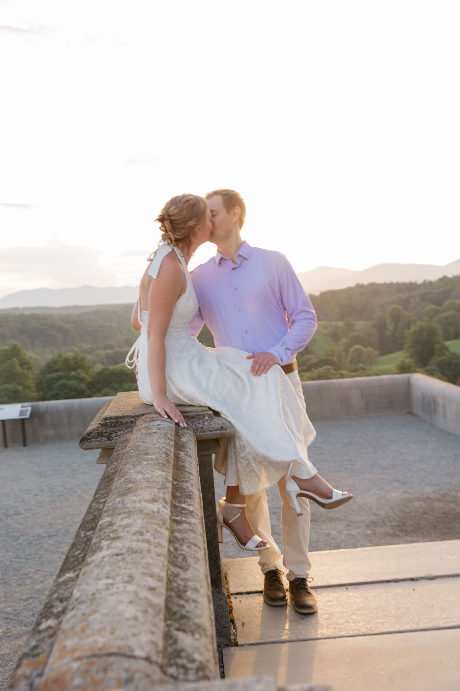 Couple sits casually on a stone wall during sunset with rolling hills in the background.