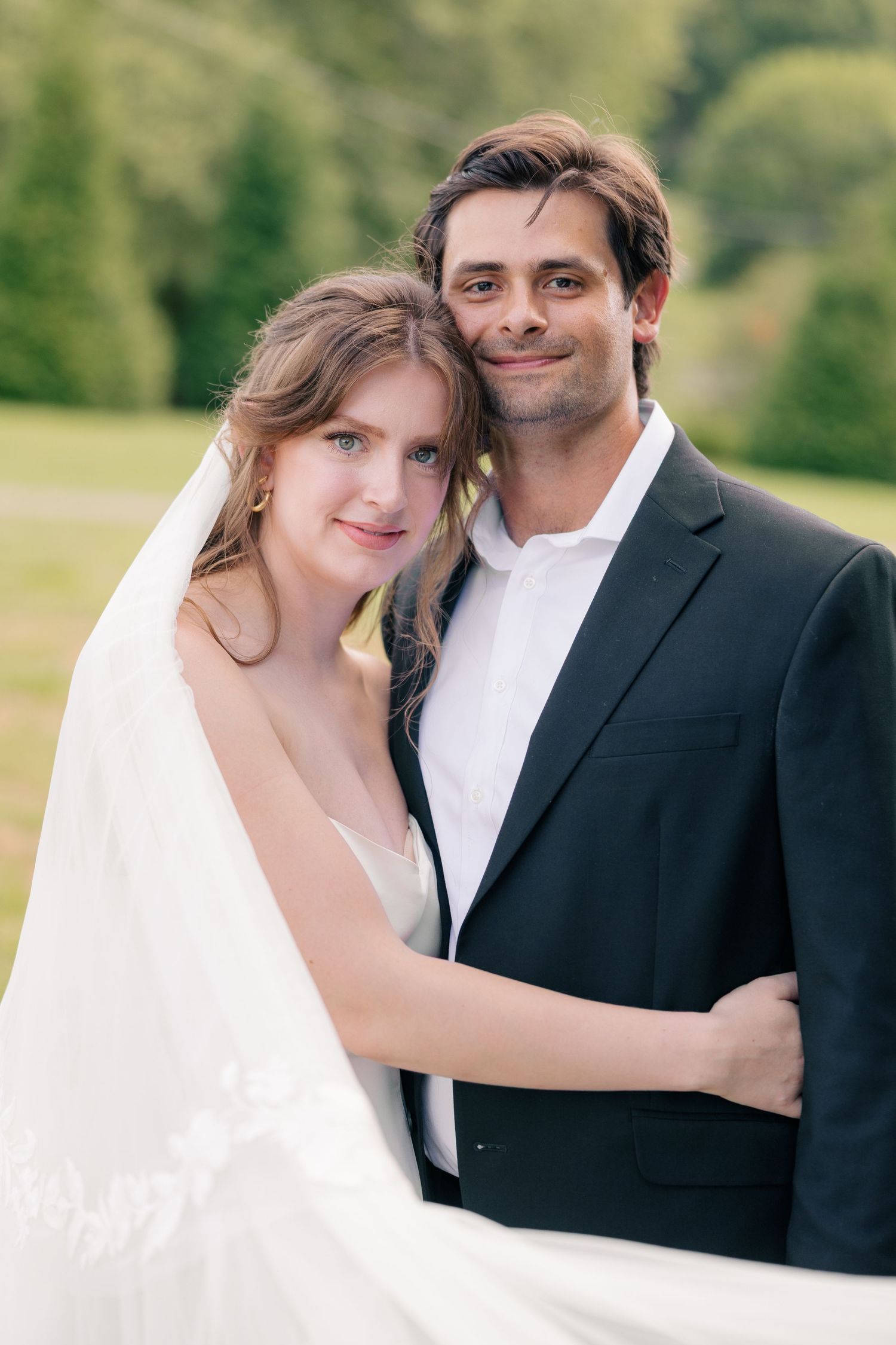 Wedding couple embracing outdoors, bride in white gown with veil and groom in dark suit smiling at camera.