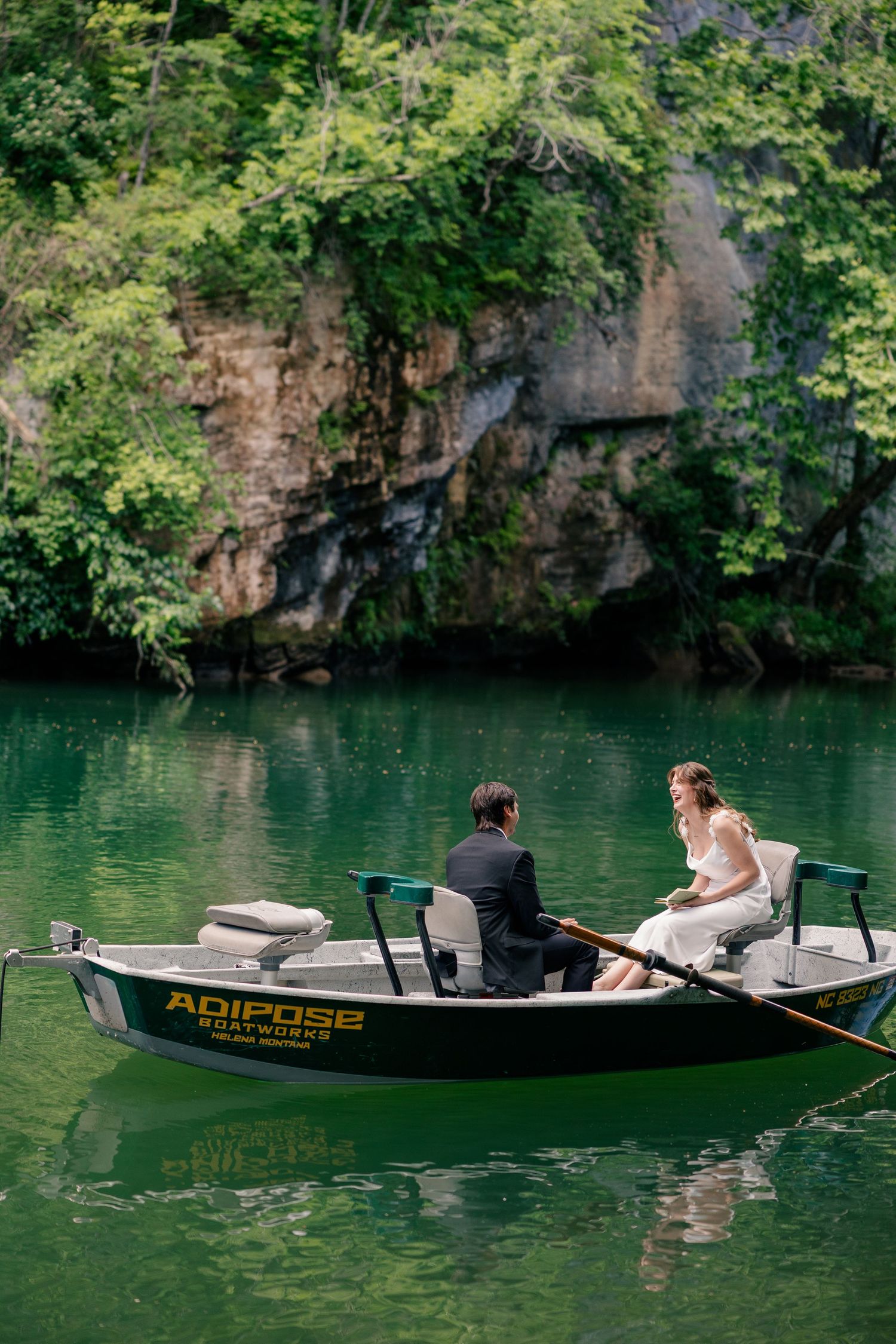 Couple in wedding attire seated in an Adipose Boatworks drift boat on a calm emerald green river.