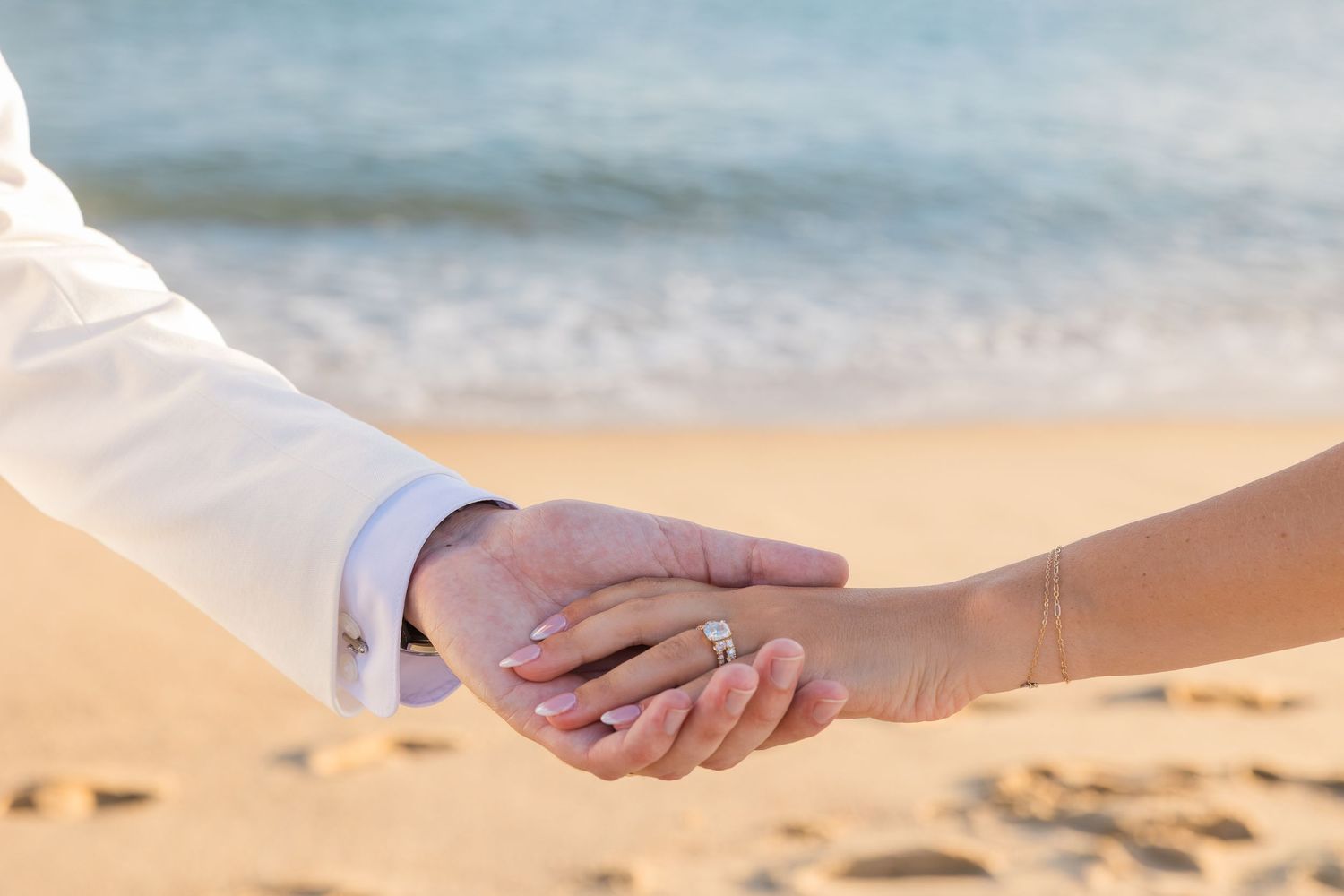 Couple holding hands on a sandy beach, bride wearing a diamond engagement ring, ocean waves in the background.